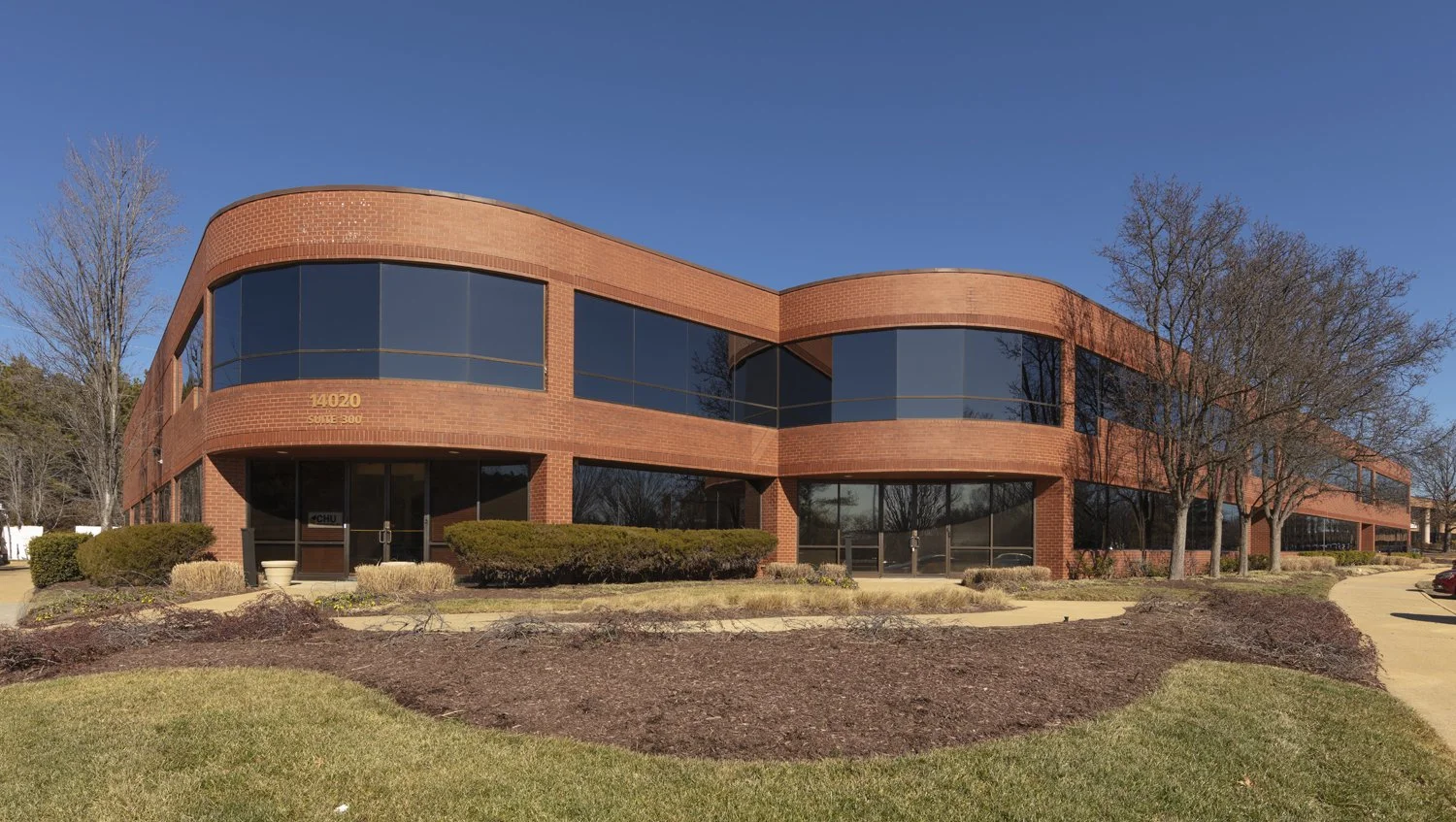 A two-story brick office building with large tinted windows, surrounded by trees, bushes, and a manicured lawn under a clear blue sky.