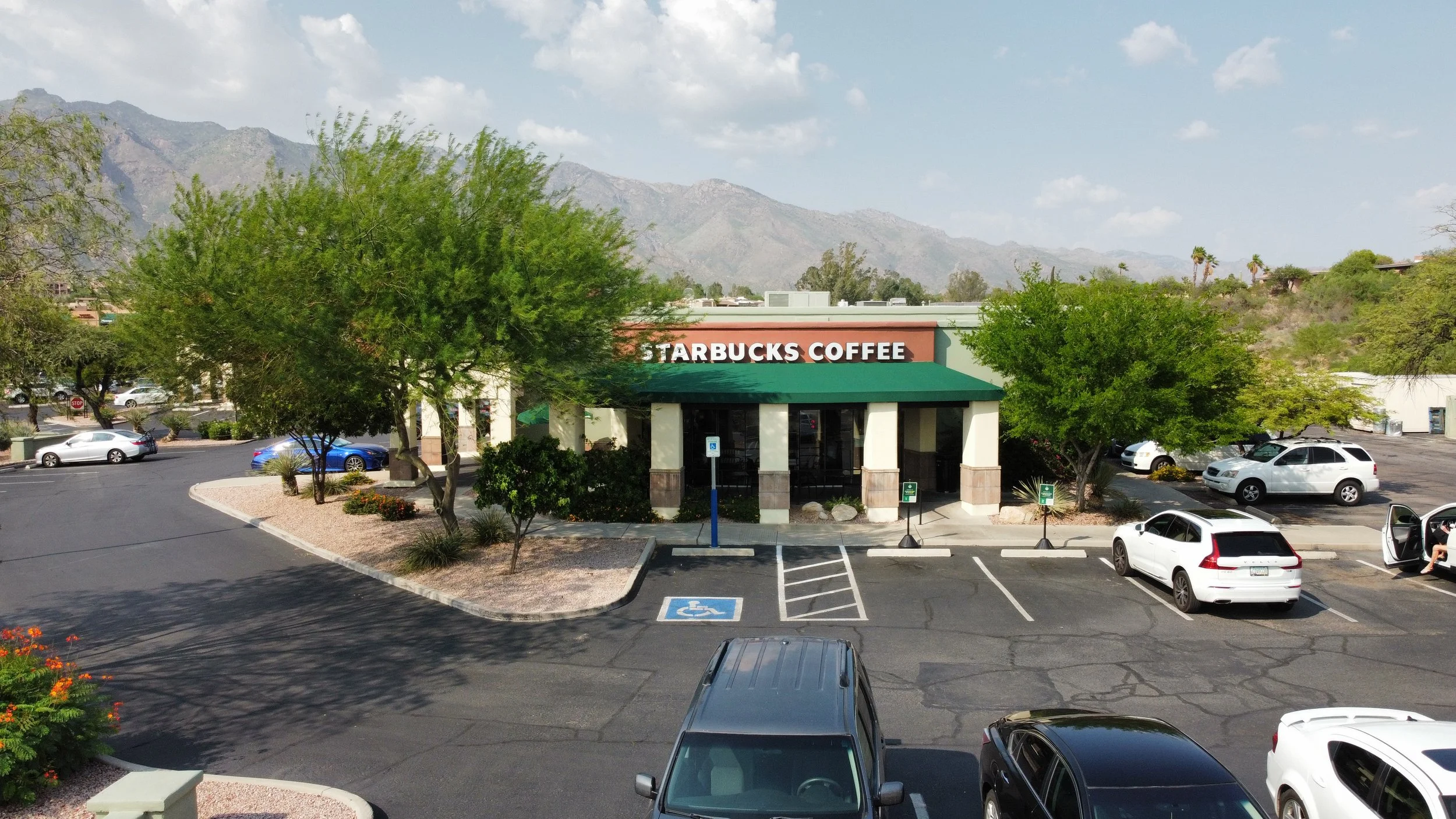 Starbucks Coffee store in a parking lot with trees, cars, mountains in the background, and a clear sky with scattered clouds.