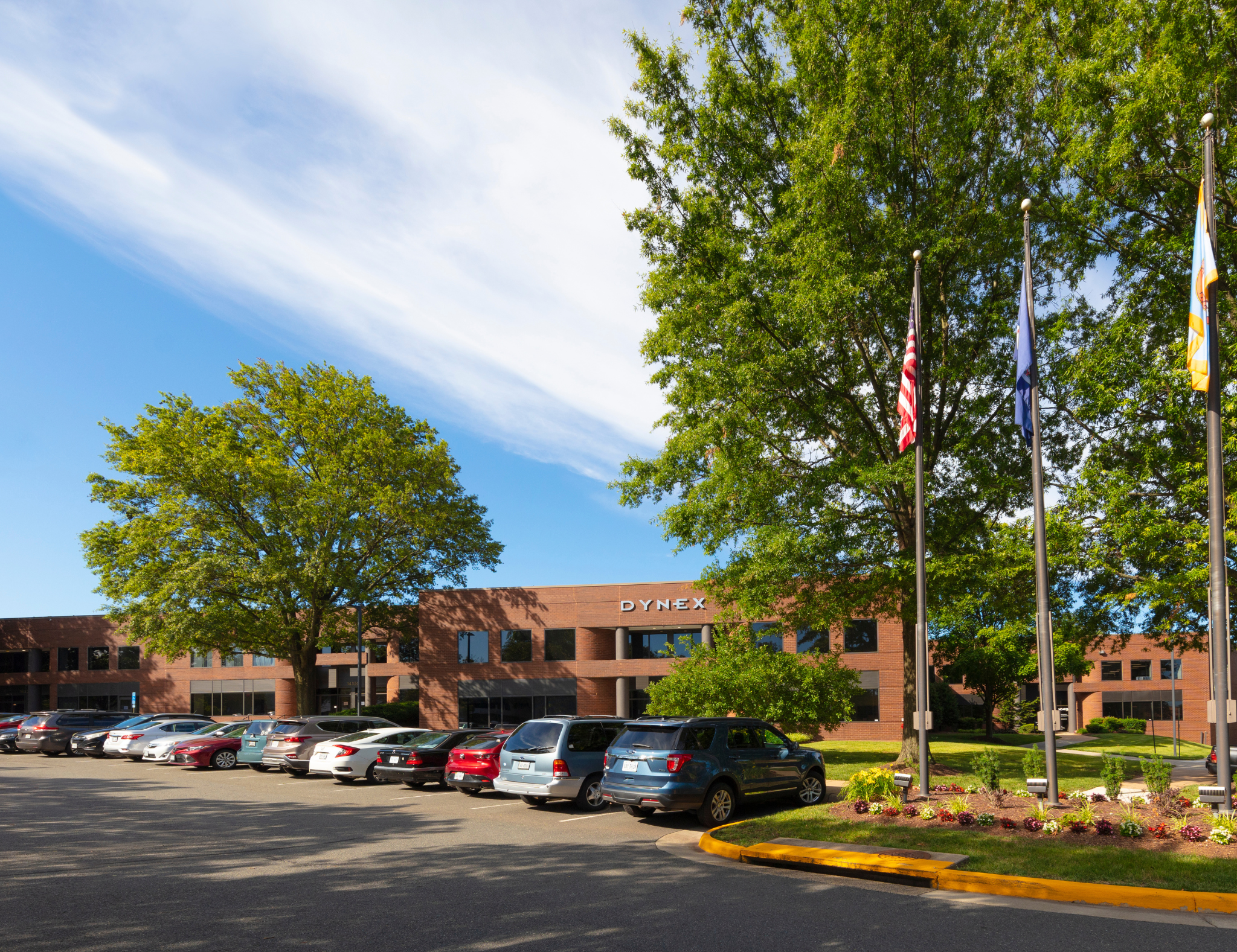 A parking lot filled with cars in front of a brick office building with the sign "Dynex." Several trees with green foliage are visible and three flagpoles with flags are in the scene. The sky is partly cloudy with patches of blue.