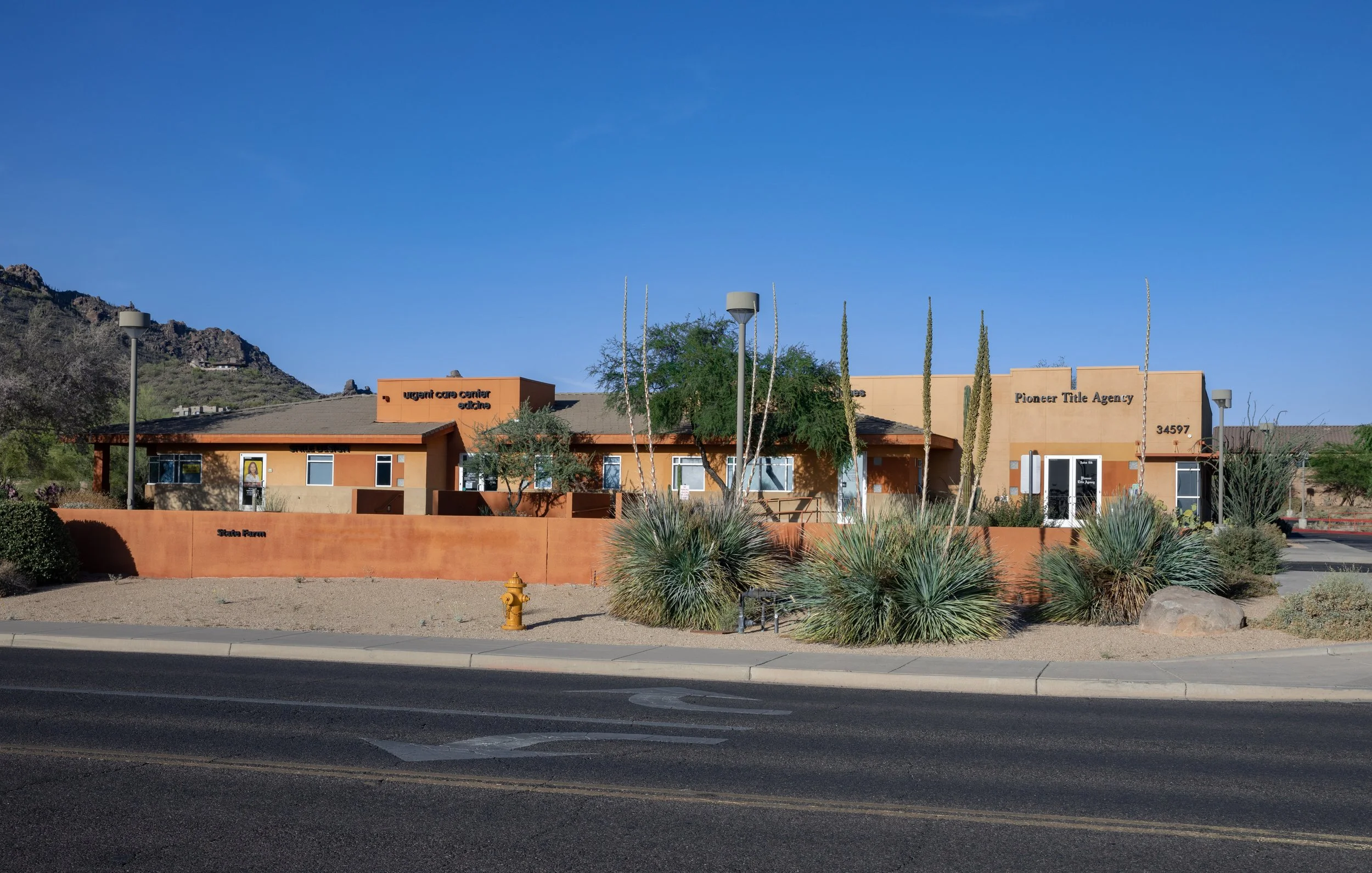 Photo of a building with a desert landscape, including cactus plants, a hill, and a bright blue sky.