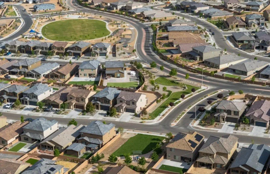Aerial view of a suburban neighborhood with rows of houses, streets, a park with a green field, and landscaped yards.
