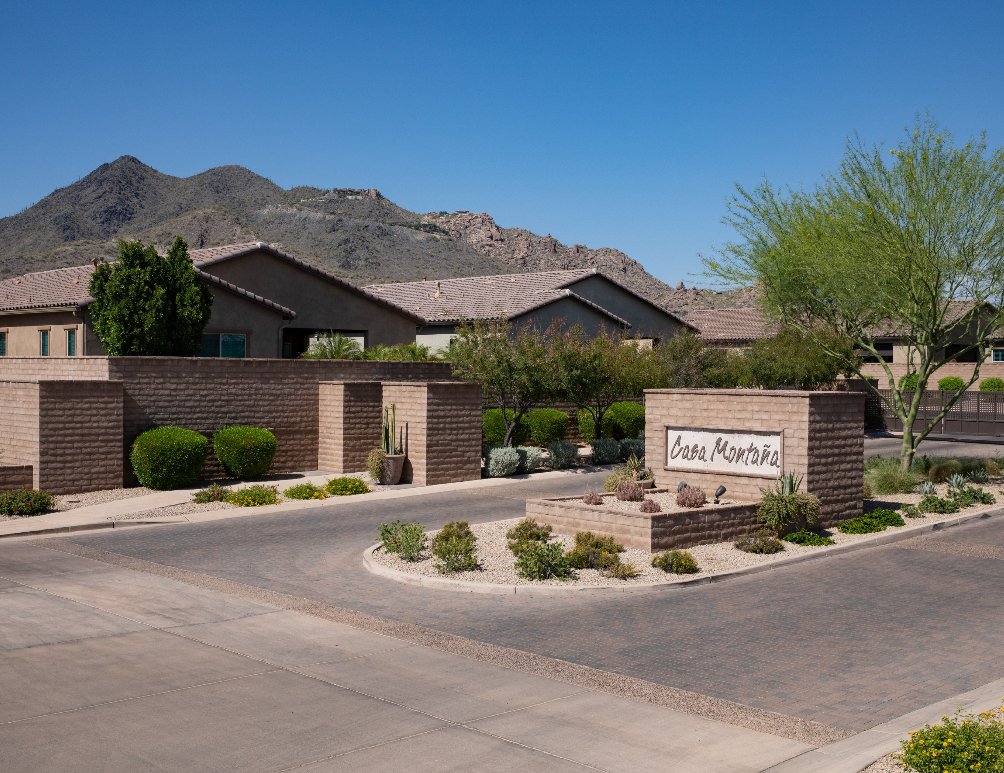 View of a residential area at the base of desert mountains under a clear blue sky. The houses have stucco walls and tile roofs, with a landscaped yard in the foreground featuring various bushes, small trees, and a sign that reads 'Casa Montana'.