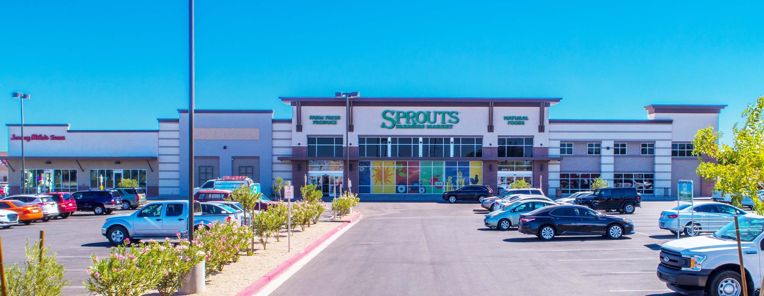 Exterior of Sprouts Farmers Market grocery store in a shopping plaza with parking lot and cars, storefront has large windows and colorful graphics.