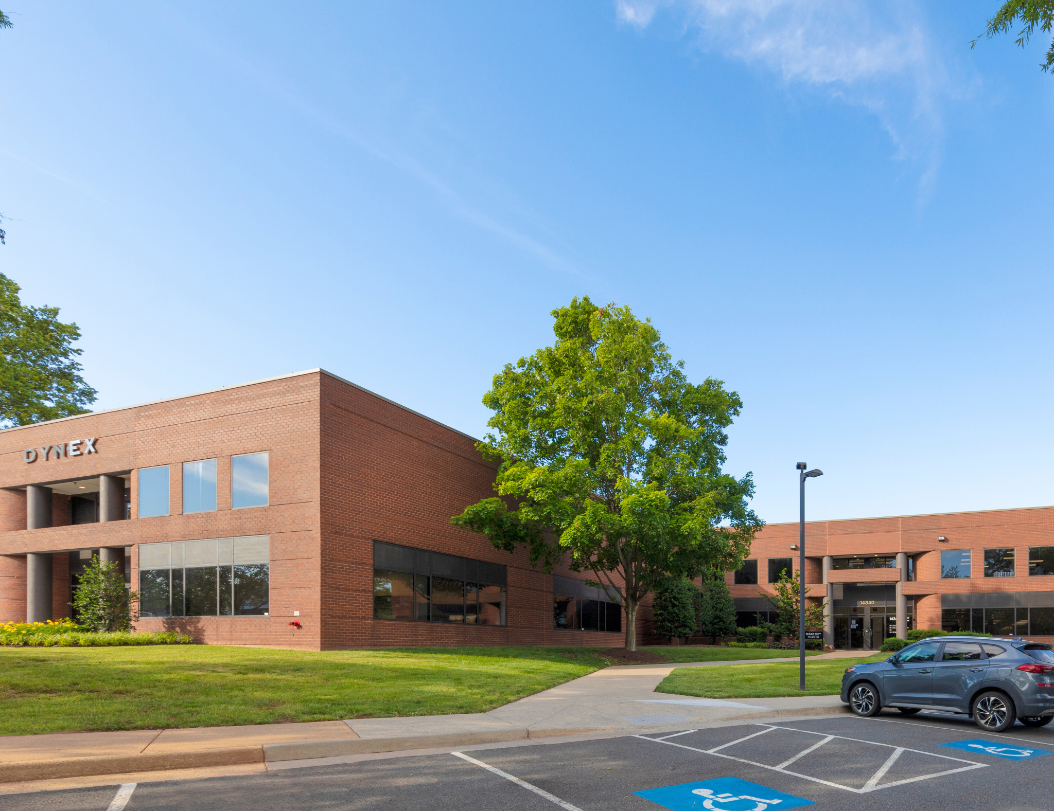 A brick office building with windows and the word "DYNEX" on it, surrounded by green trees and a parking lot with a blue handicapped parking space.
