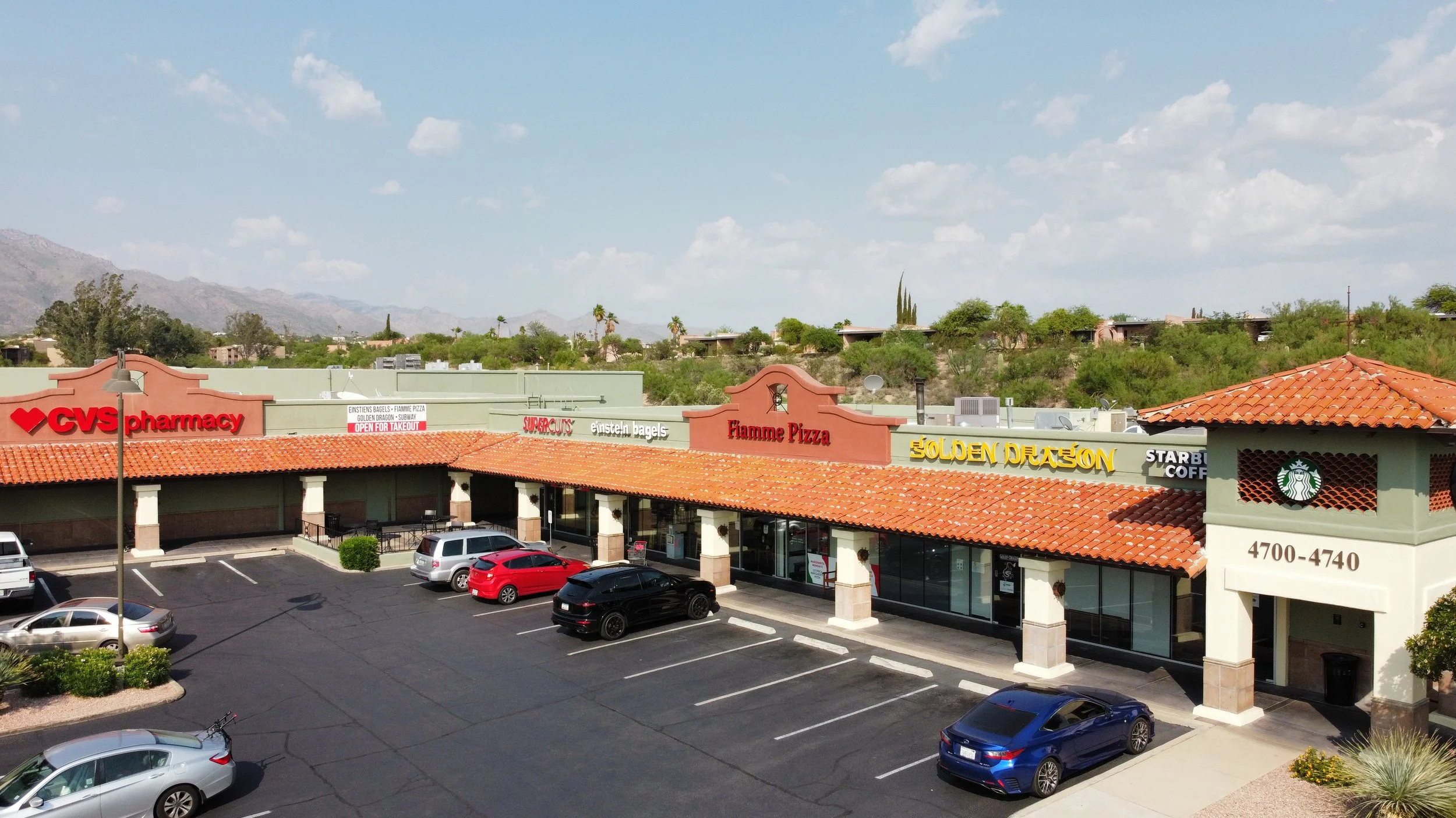 Shopping plaza with storefront signs including CVS Pharmacy, Einstein Bagels, Fiamme Pizza, Golden Dragon, Starbucks Coffee, and Supercuts, parking lot with several cars, and mountain and desert landscape with sparse trees in the background.