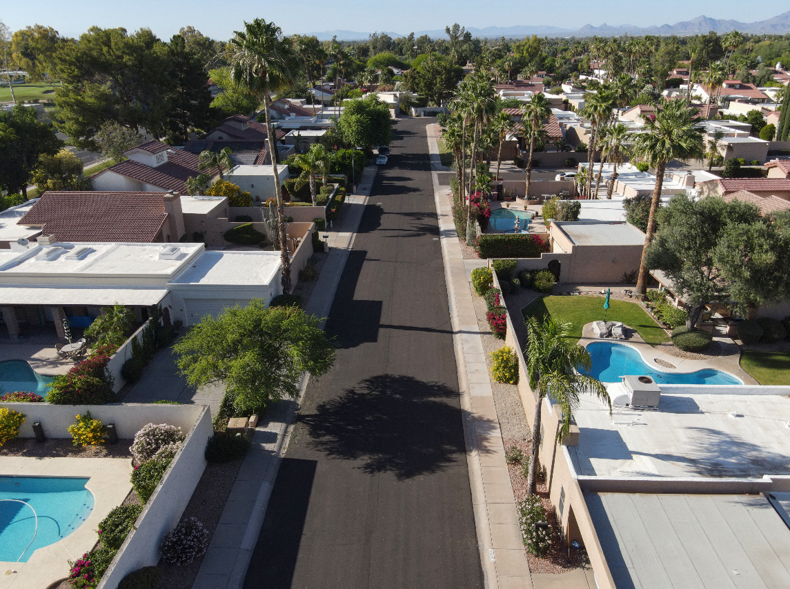 Aerial view of a suburban neighborhood with single-story houses, palm trees, and backyard swimming pools, with mountains in the background.