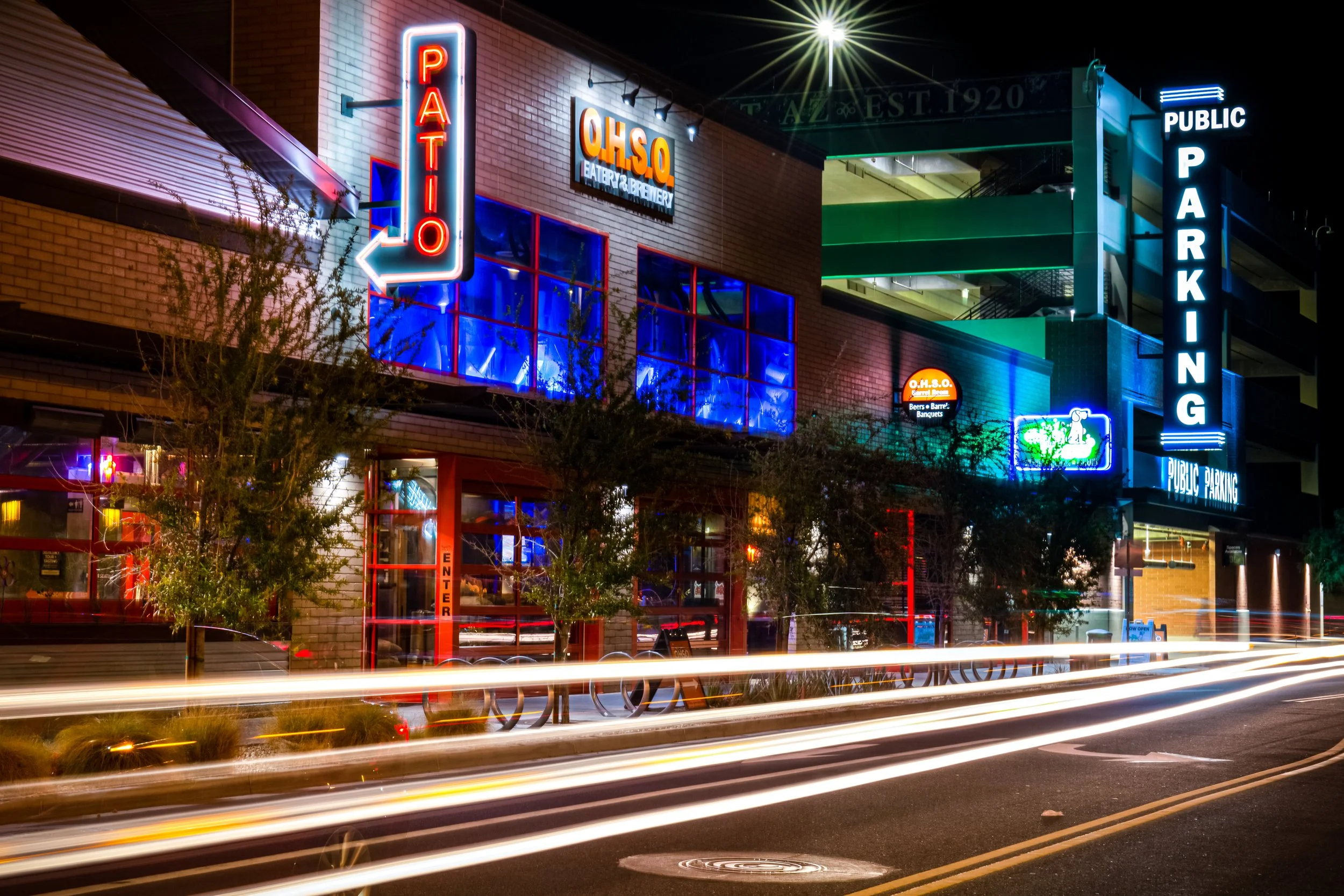 Nighttime street scene with illuminated neon signs for a parking garage, a restaurant called OHSO with blue and orange lighting, and other storefront signs. Light streaks from moving vehicles are visible in the foreground.