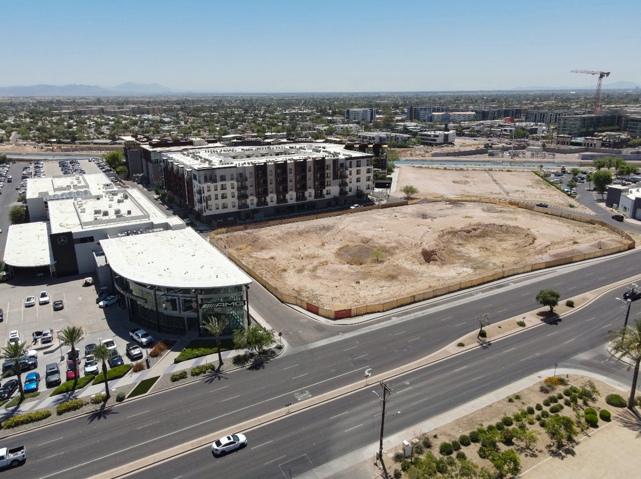 Empty lot of dirt and sand is surrounded by fencing, located next to a dealership with a Mercedes-Benz logo, near a new residential building, with a parking lot and cars nearby, under a clear blue sky.