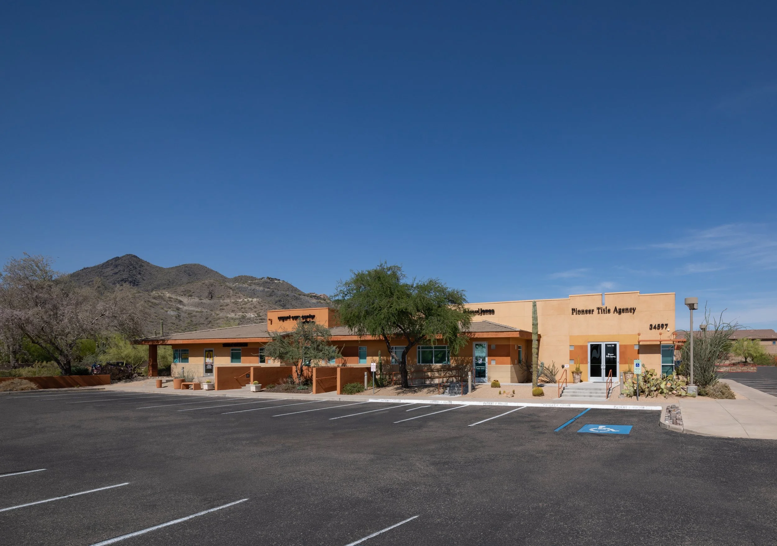A single-story building with a sign that reads 'Pioneer Title Agency' situated under cloudy skies, with mountains in the background, a parking lot with several empty spaces, including a handicapped parking spot, and desert vegetation around the building.