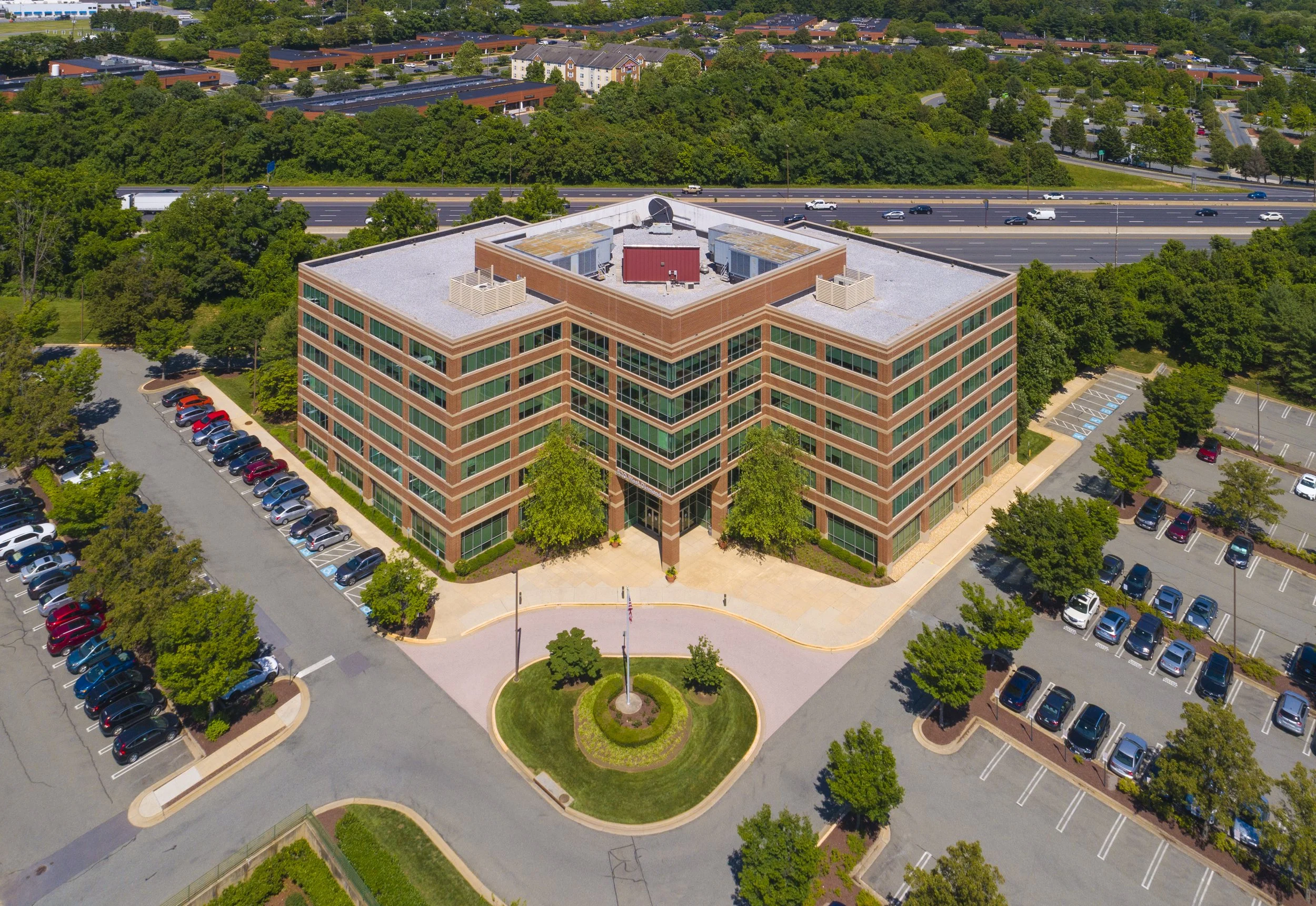 Aerial view of a multi-story office building with parking lots and trees surrounding it, and a highway in the background.