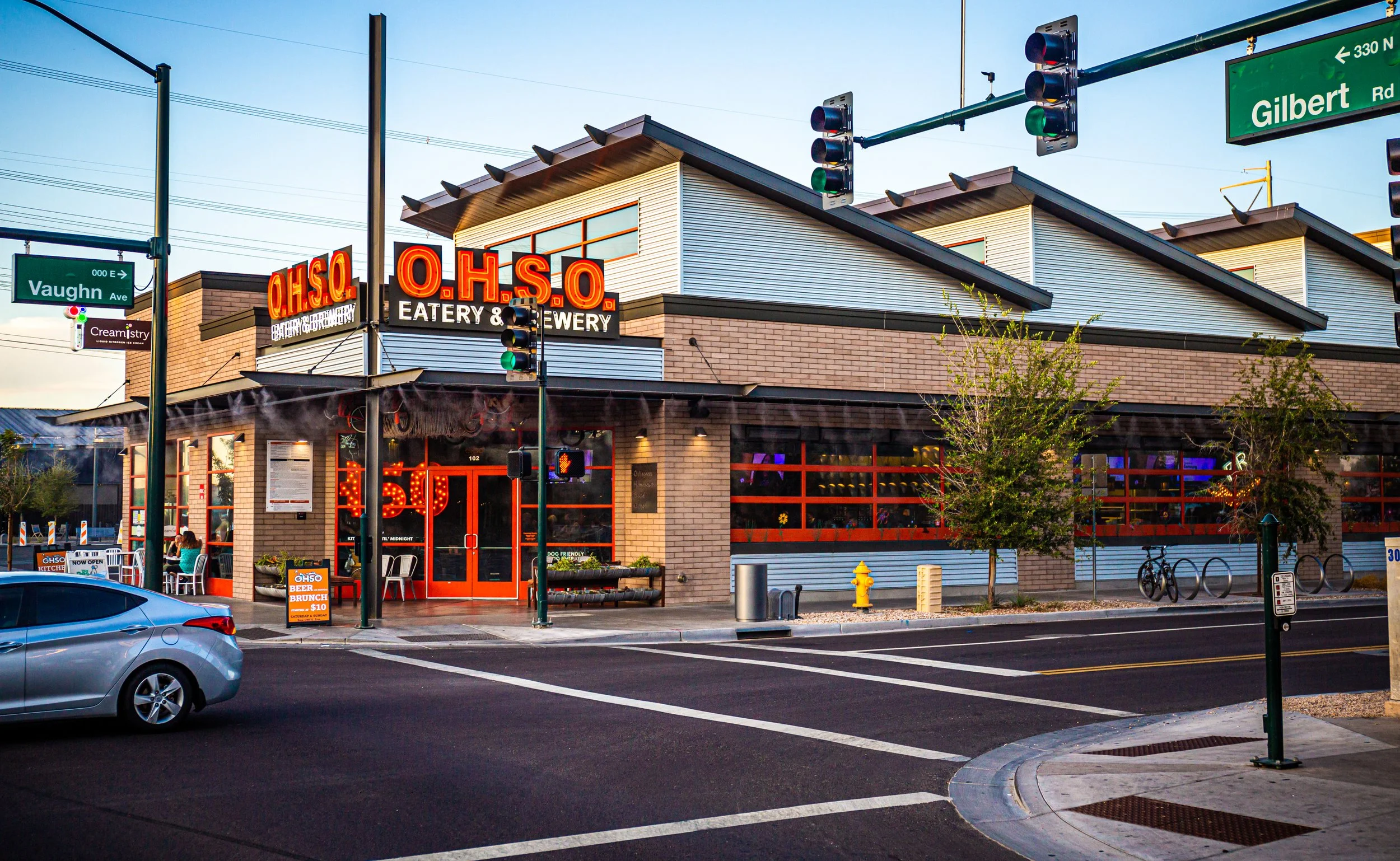 A modern restaurant building with a neon sign that reads "O.H.S.O. Eatery & Brewery." The restaurant features brick and siding exterior, large windows, and outdoor seating. Traffic lights are visible at the intersection, along with street signs for Vaughan Avenue and Gilbert Road. The scene appears to be in the early evening with clear skies.