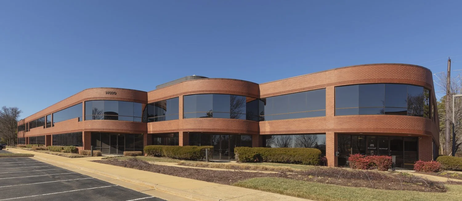 A two-story office building with brick exterior and large reflective windows, surrounded by a parking lot, shrubs, and a clear blue sky.