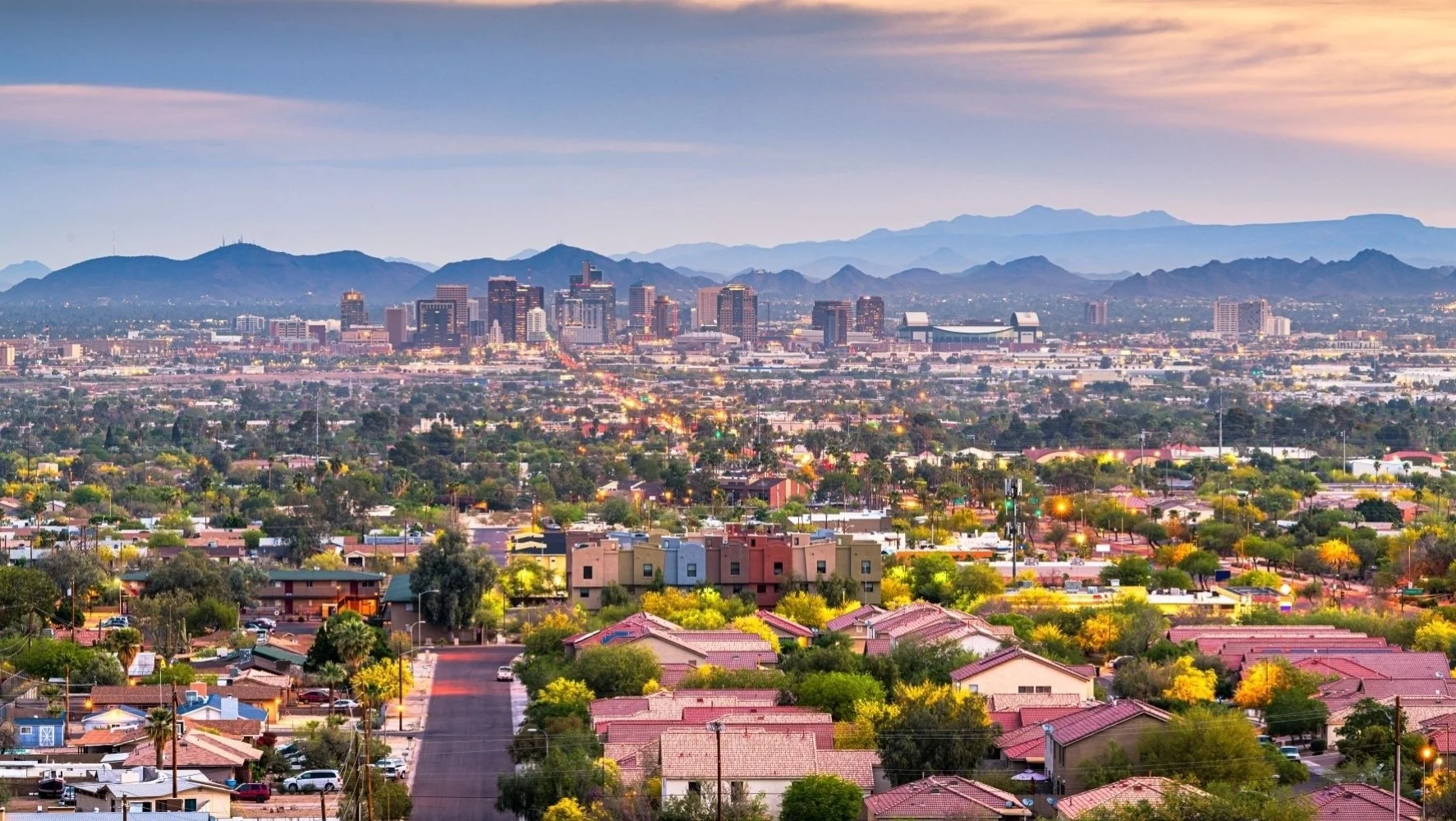 Cityscape of downtown with high-rise buildings against mountains during dusk, foreground shows residential neighborhood with streets and trees.