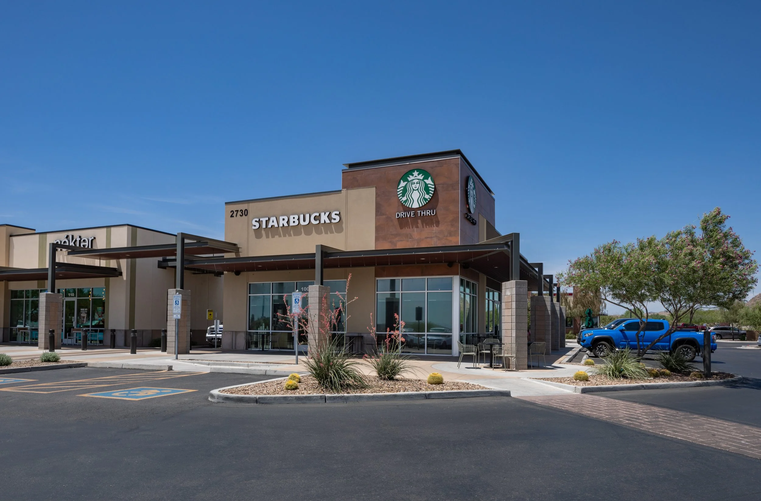 A shopping center with a Starbucks coffee shop featuring a drive-thru. The parking lot has a few cars, including a blue truck, and there are landscaped areas with small plants and trees.