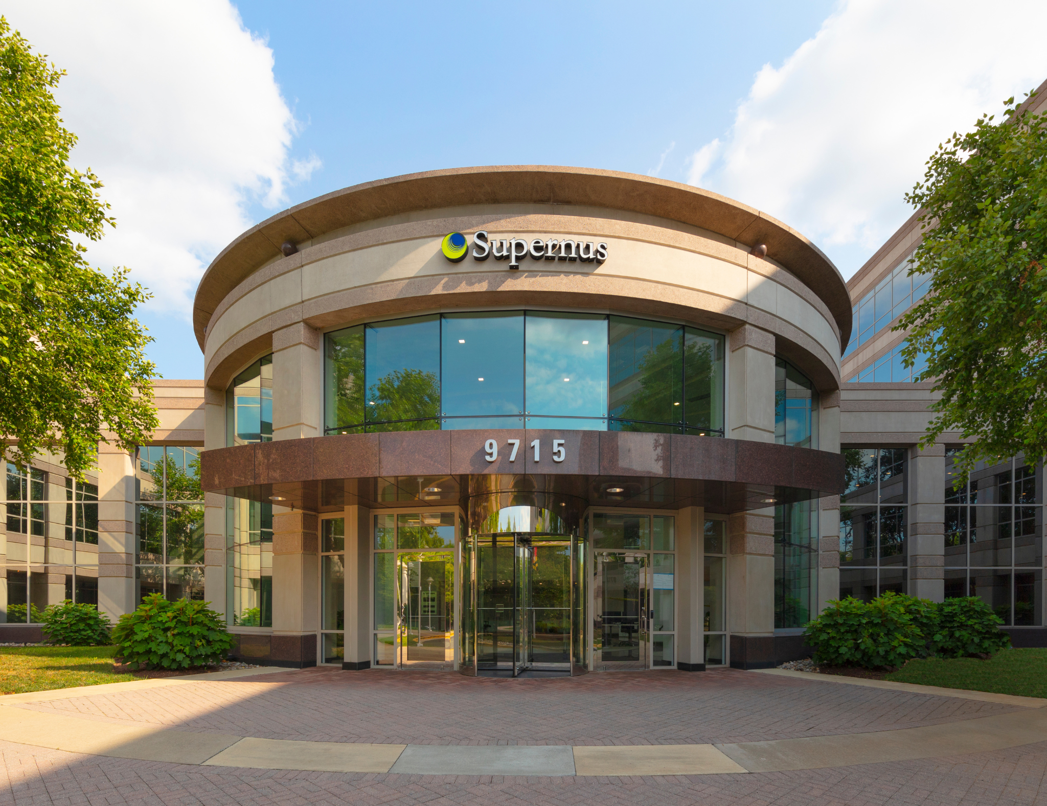 Modern office building with curved glass entrance, green trees, and a partly cloudy sky.