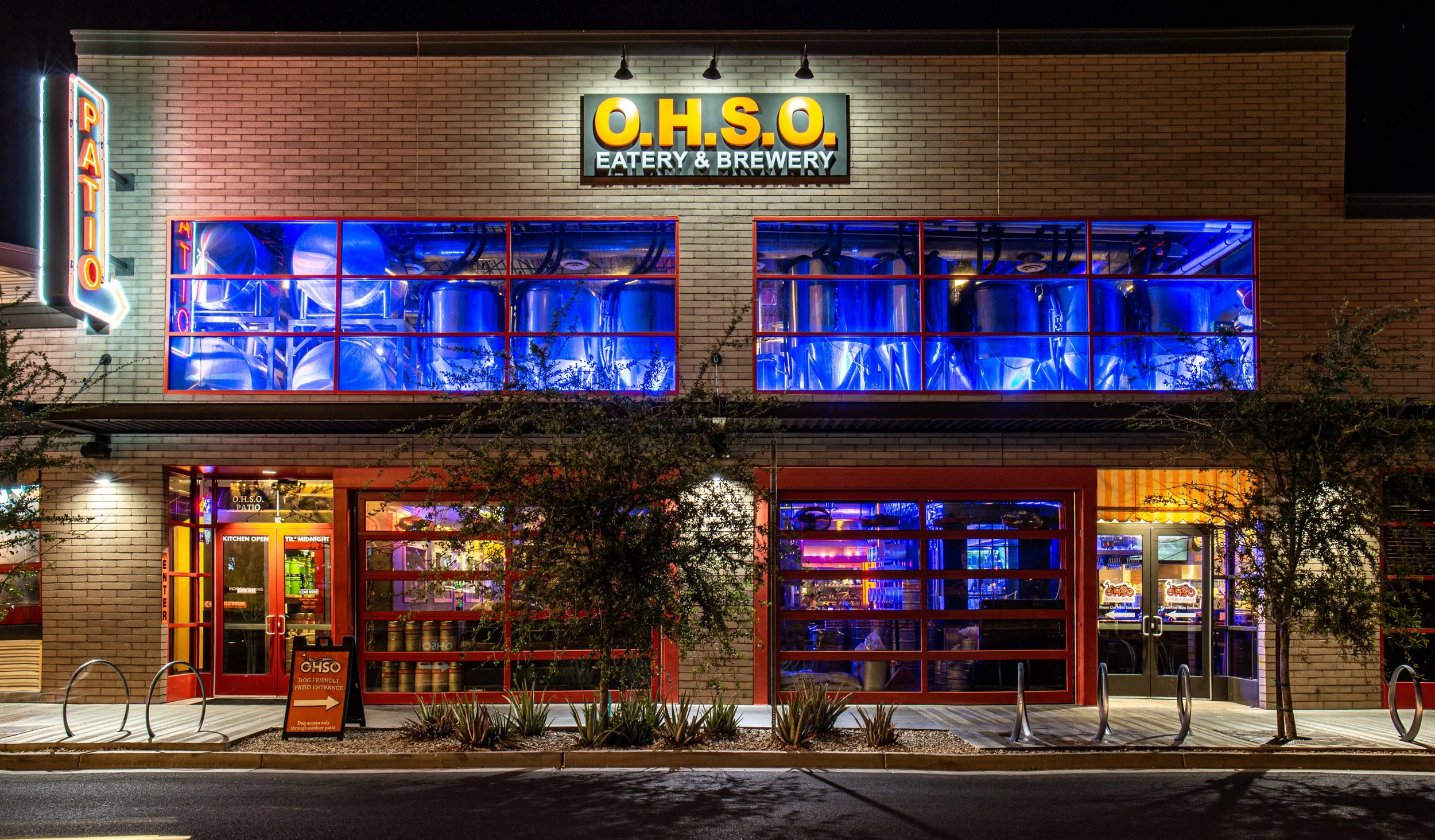Night view of O.H.S.O. eatery and brewery with illuminated sign, large windows showing inside taproom, and a neon patio sign. The building has a brick facade with colorful lighting and a patio area in front.