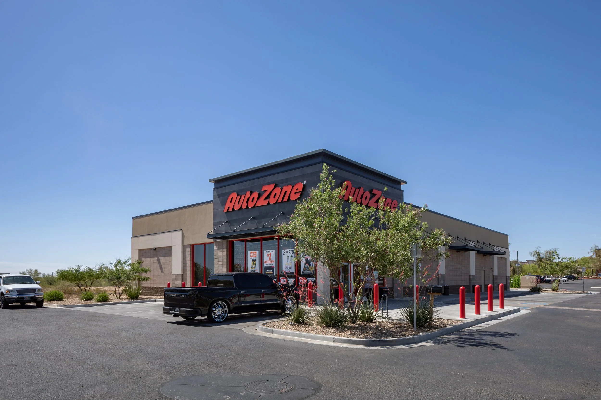 AutoZone store in a shopping center with cars parked in the lot, clear blue sky, and a few trees in front of the store.