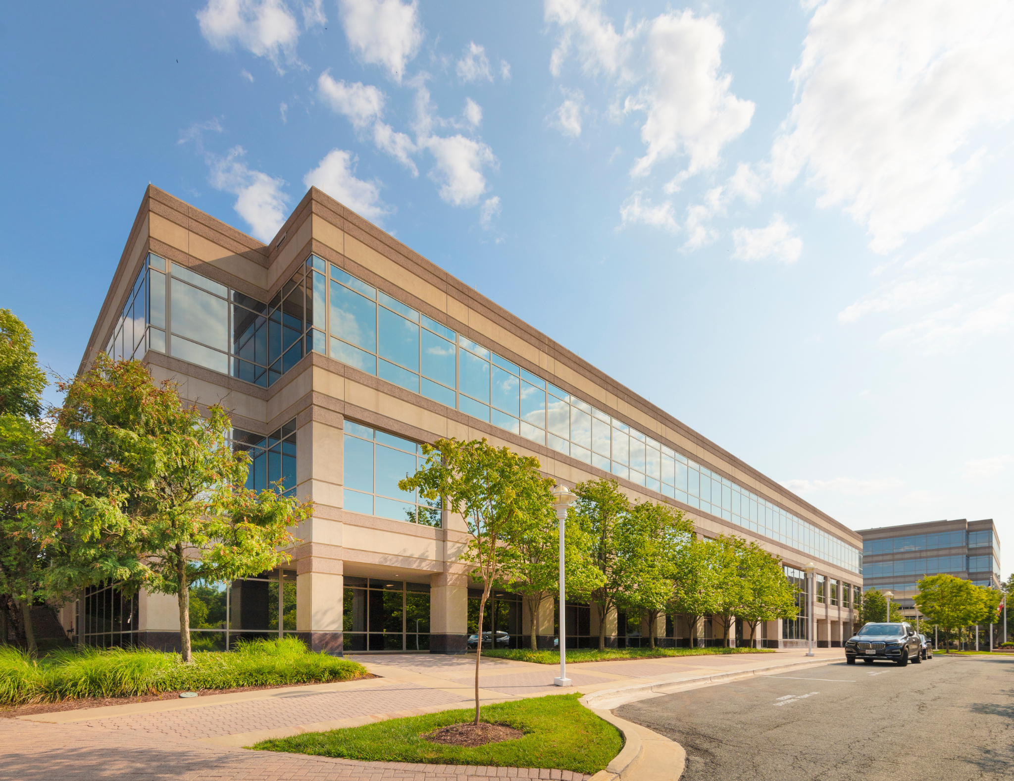 A modern office building with glass windows and beige exterior walls, surrounded by green trees and a sidewalk, under a partly cloudy blue sky.