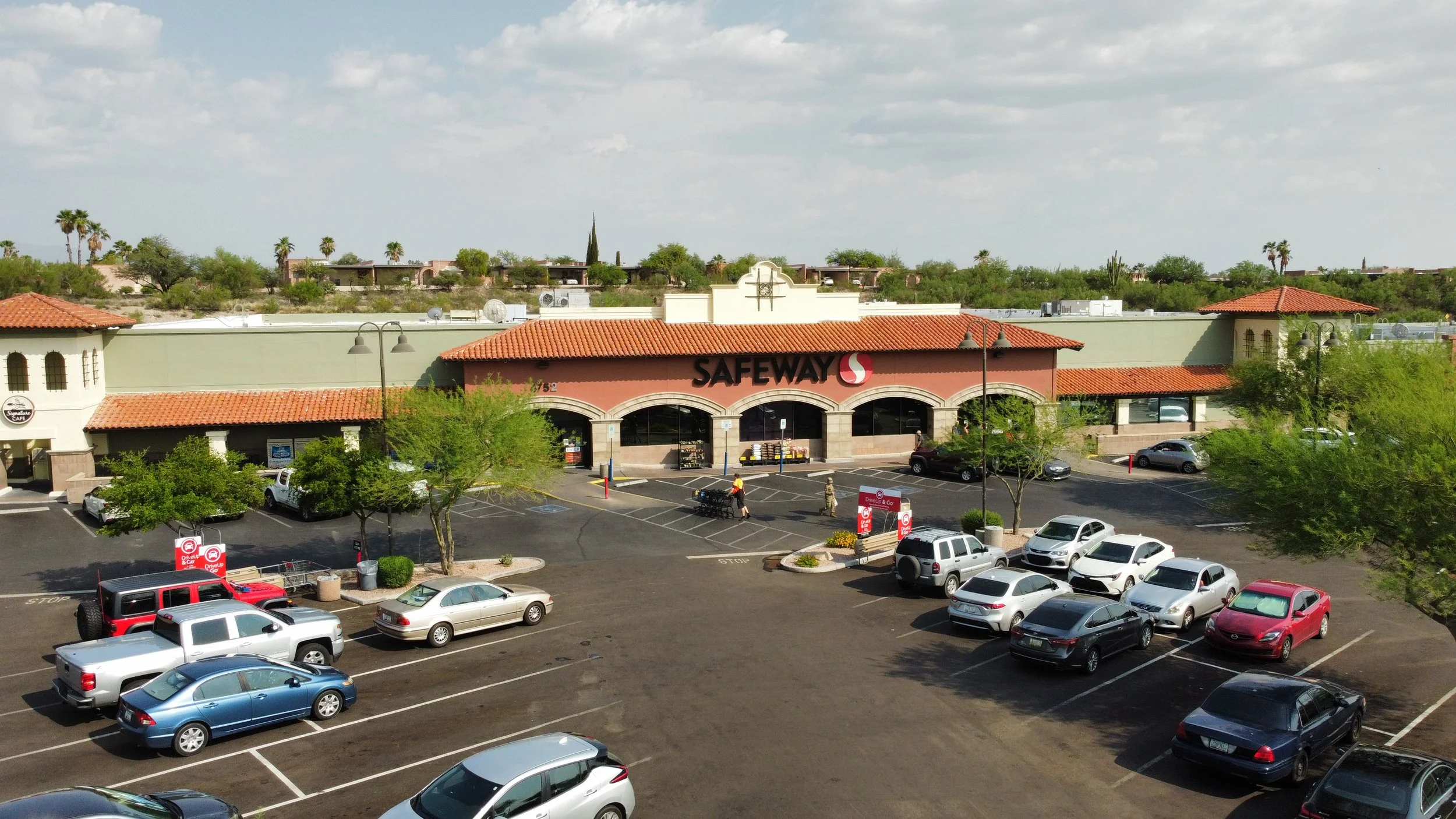 Parking lot view of Safeway supermarket with cars parked and shopping carts, desert landscaping, trees, and buildings in the background under a partly cloudy sky.