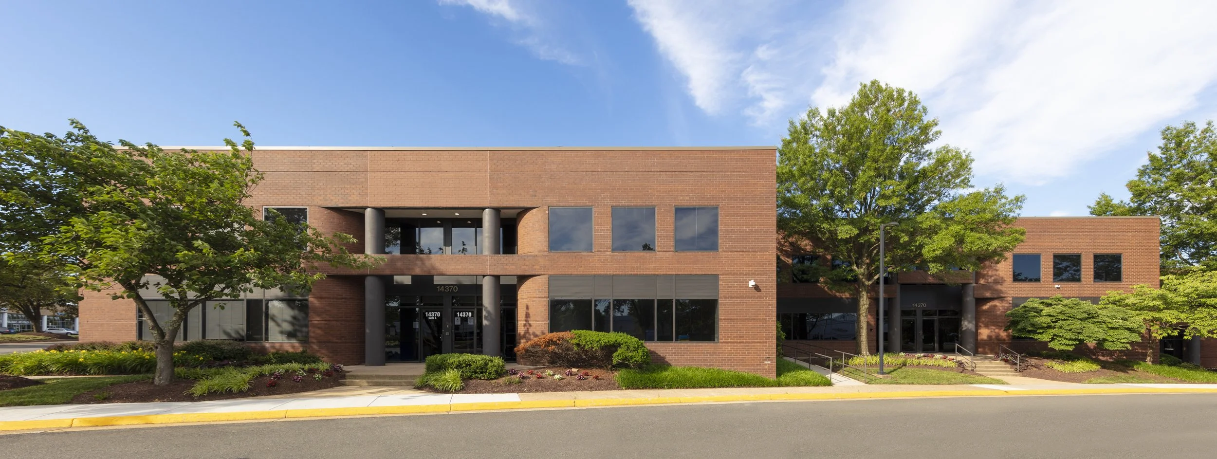 A modern two-story brick office building with large windows, surrounded by trees and landscaped greenery, under a partly cloudy sky.