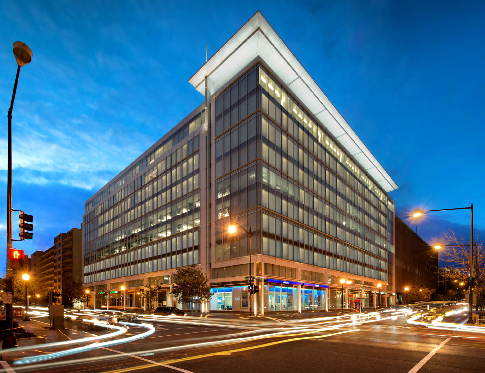 Modern office building with glass windows at twilight, with light trails from passing cars in the foreground and streetlights illuminating the scene.