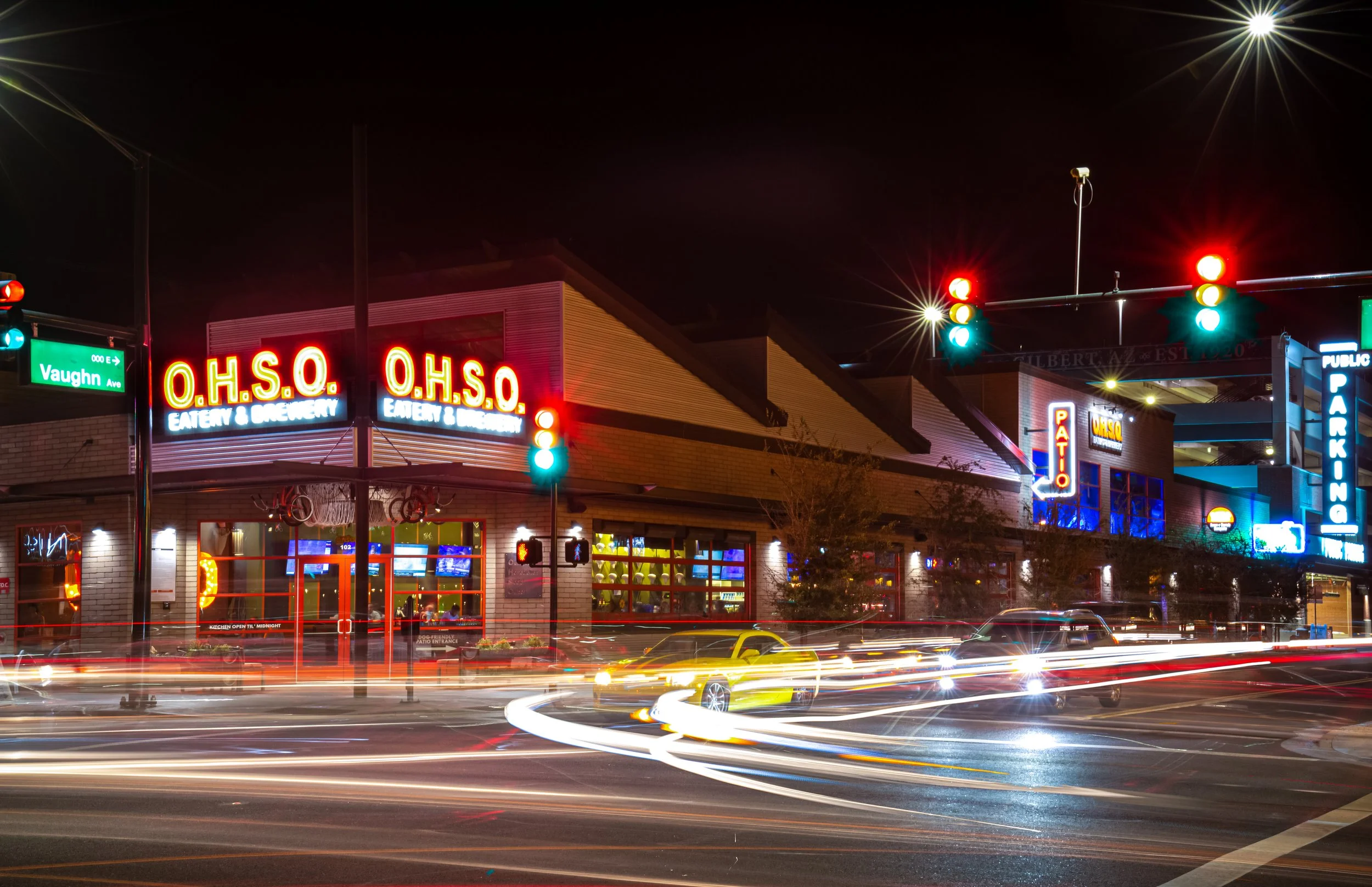 Night view of a busy street with a restaurant named O.H.S.O. Eatery & Brewery, illuminated red and yellow neon signs. Traffic lights show red and green signals. Light trails from moving vehicles are visible due to long exposure. The scene captures city life at night with illuminated storefronts and passing cars.