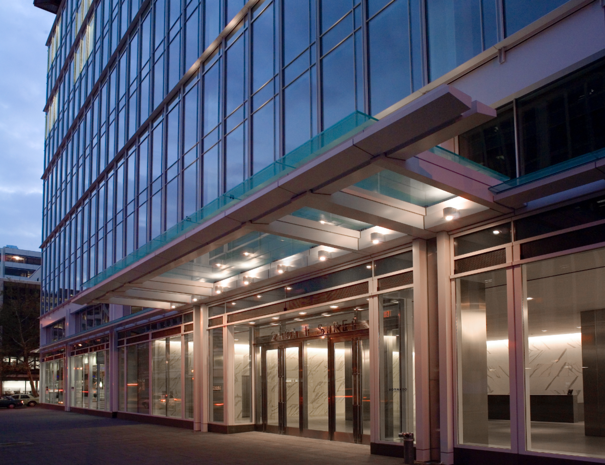 The exterior of a modern glass office building at dusk illuminated by interior and exterior lights, with a parking lot in front.