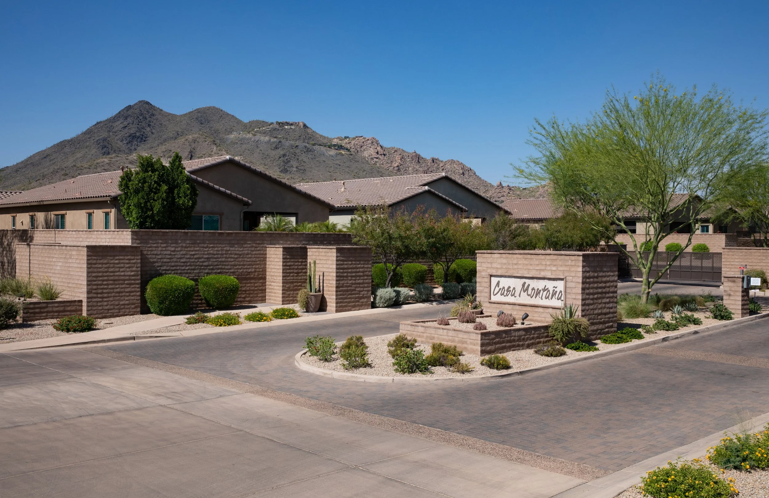 A residential gated community entrance with a beige brick wall and a sign that reads 'Casa Montana'. There are well-maintained bushes and trees, and mountains are visible in the background under a clear blue sky.