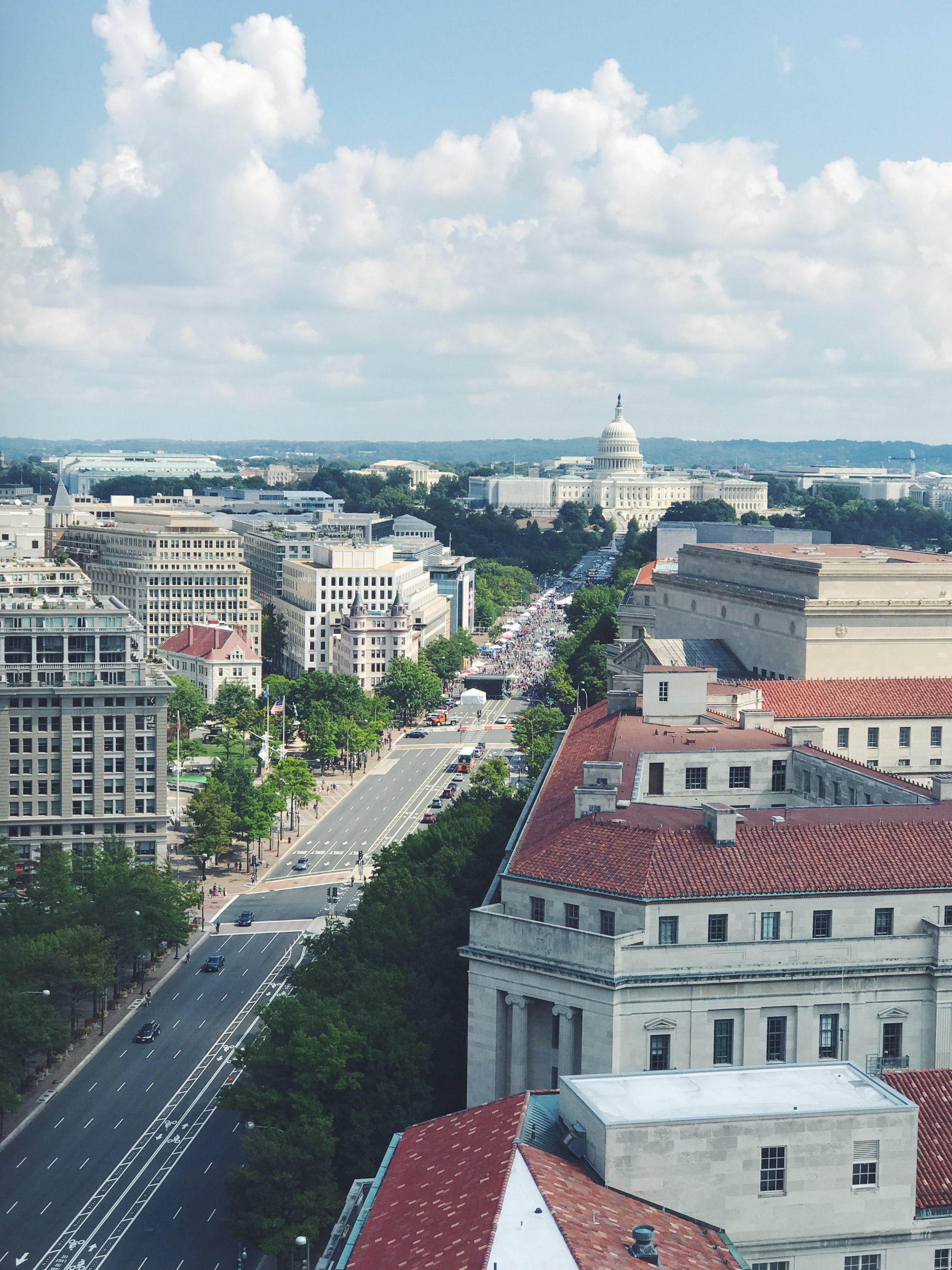 A cityscape featuring the United States Capitol building in Washington, D.C., with a wide street lined with trees in the foreground and a partly cloudy sky overhead.