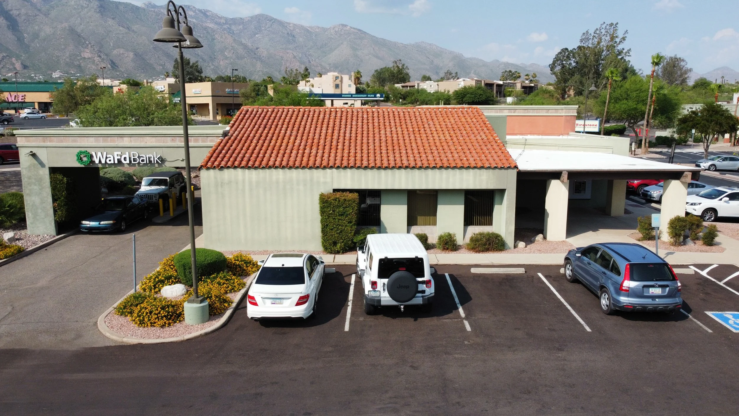 Parking lot in front of a bank building with a red tile roof, surrounded by cars and bushes, with mountains in the background.