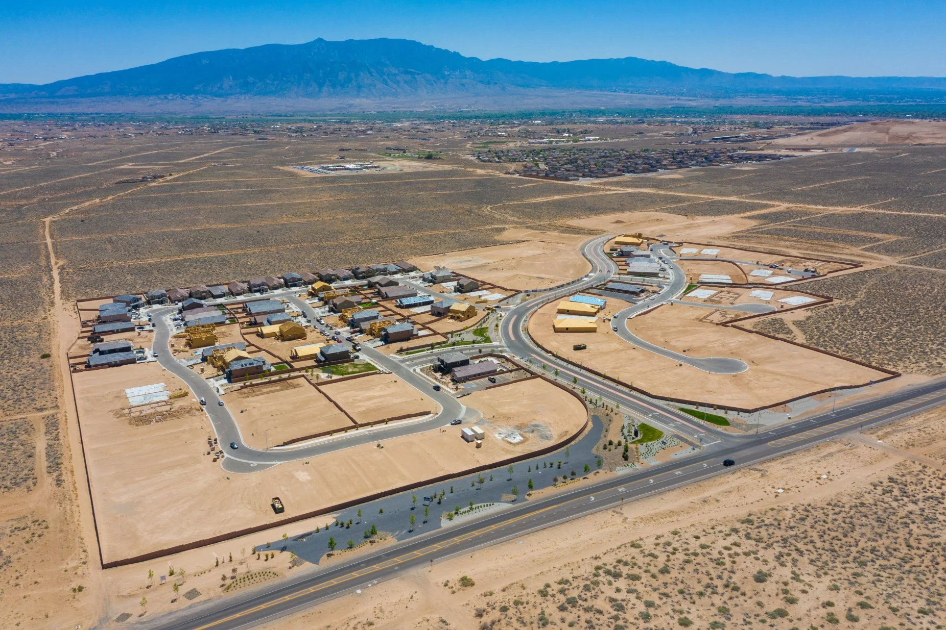 Aerial view of a new residential neighborhood under development in a desert landscape with mountains in the background. The neighborhood features houses, roads, and green spaces, with some homes still under construction.