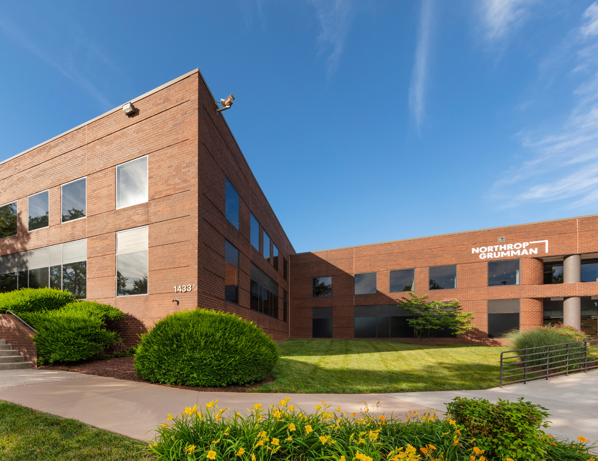 A brick office building with multiple windows and a sign that reads 'Northrop Grumman' on the front. The building is surrounded by neatly trimmed bushes and a sidewalk, with a clear blue sky overhead.