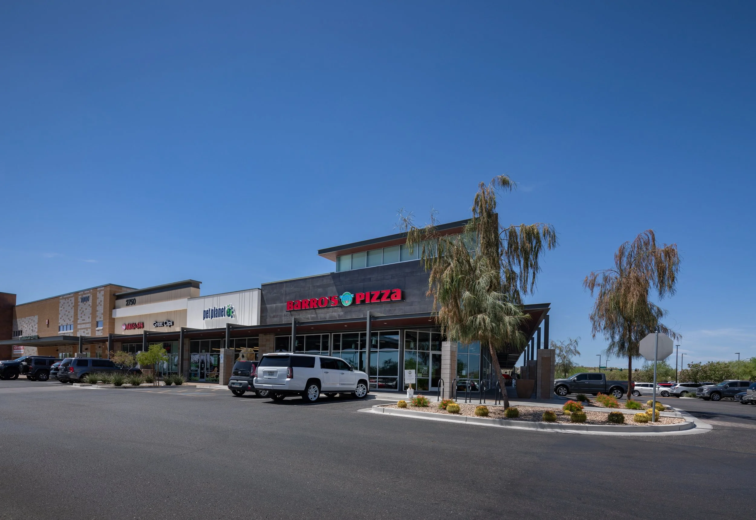 A shopping plaza with stores, parked cars, and a clear blue sky. The storefronts include Barro's Pizza, pet store, and a nail salon, with a landscaped parking lot in front.