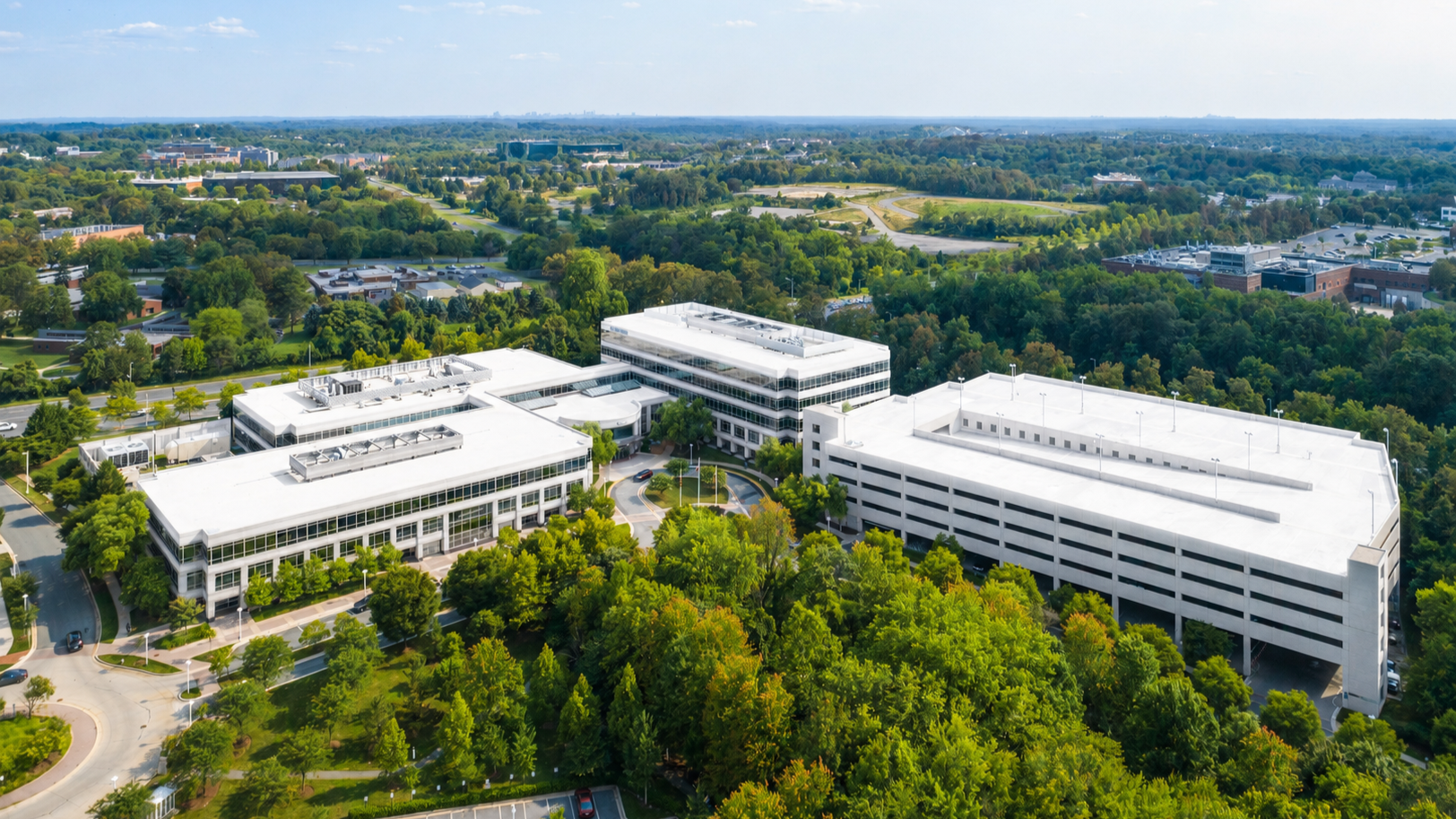 Aerial view of a corporate office complex with multiple white buildings surrounded by lush green trees and parking lots, in a suburban area with dense forest in the background.