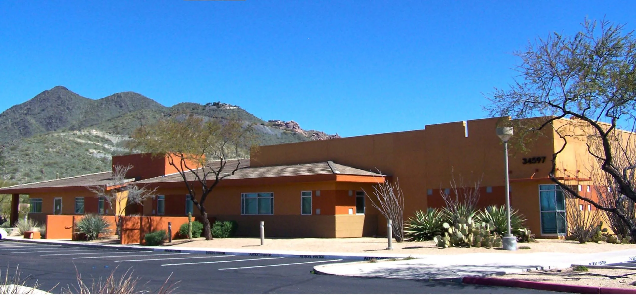 A single-story building with an orange and yellow exterior, surrounded by desert plants and trees, with a mountain range in the background under a clear blue sky.