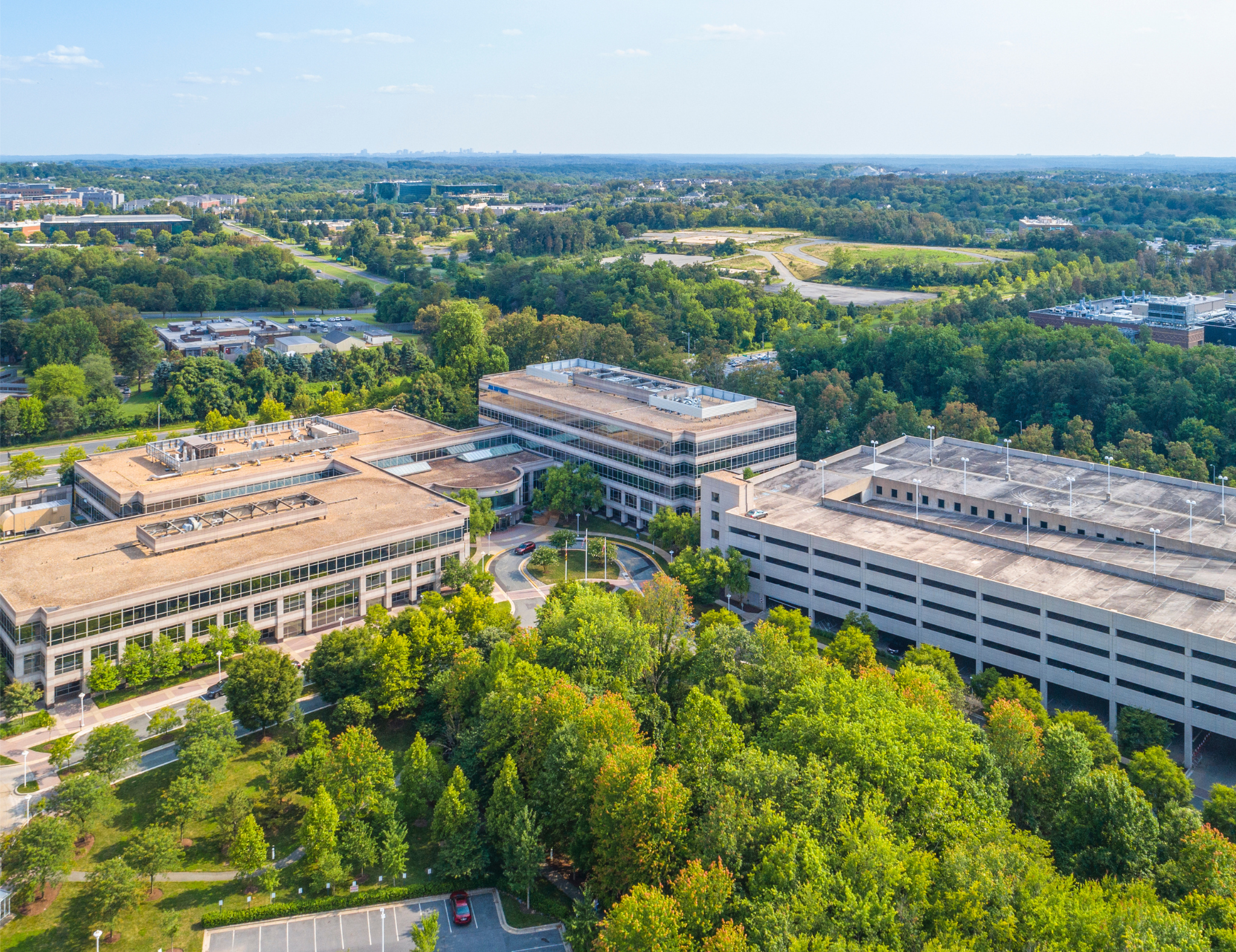 A cityscape view showing several office buildings, parking lots, and roads surrounded by dense green trees under a blue sky with a few clouds.