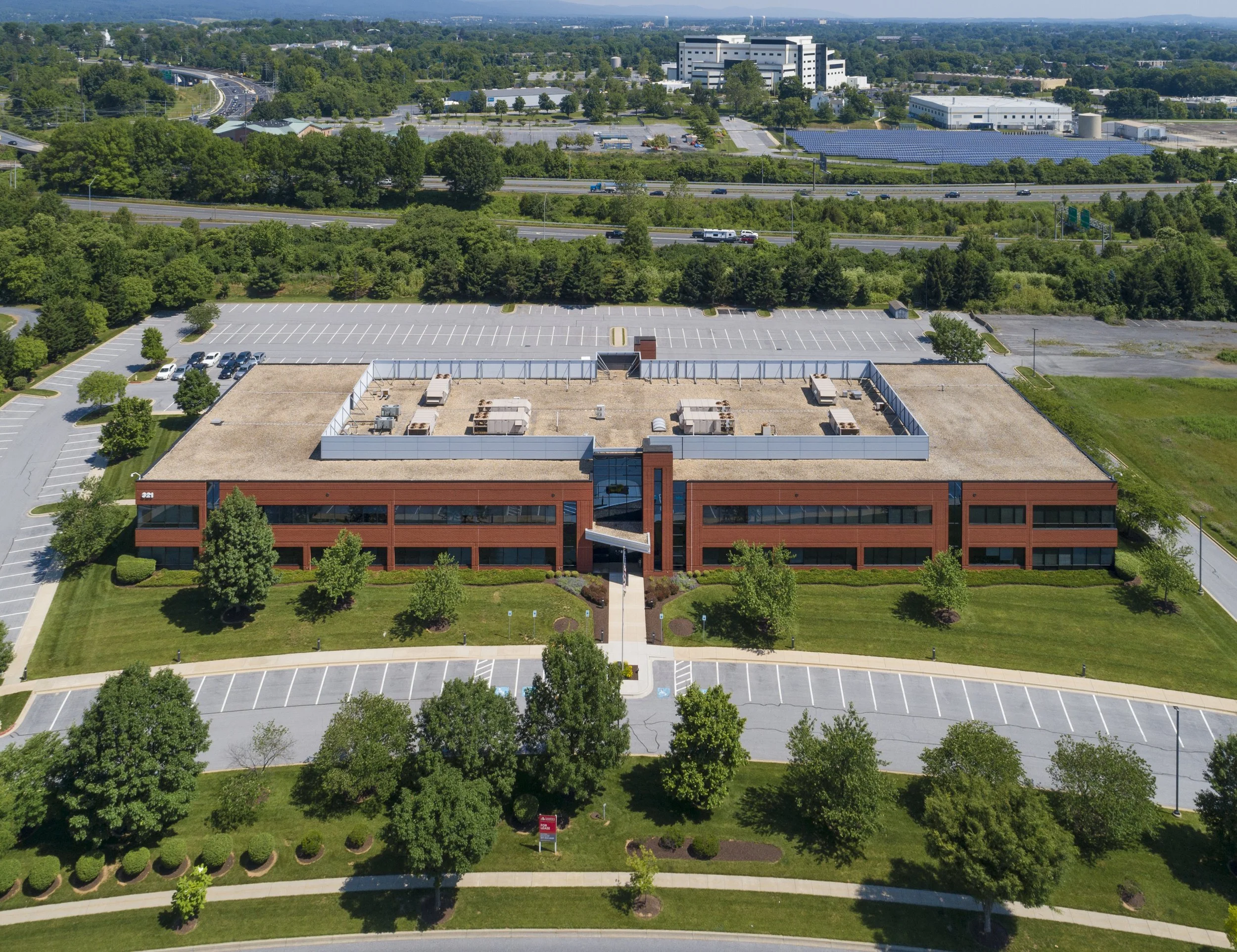 Aerial view of a large building with a rooftop parking lot, surrounded by trees and multiple empty parking lots, with a busy highway and green landscape in the background.