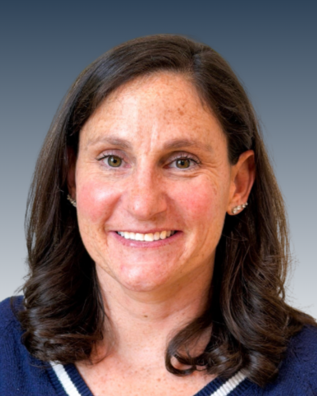 Headshot of a smiling woman with brown hair, wearing earrings and a dark-colored top, against a blurred blue background.