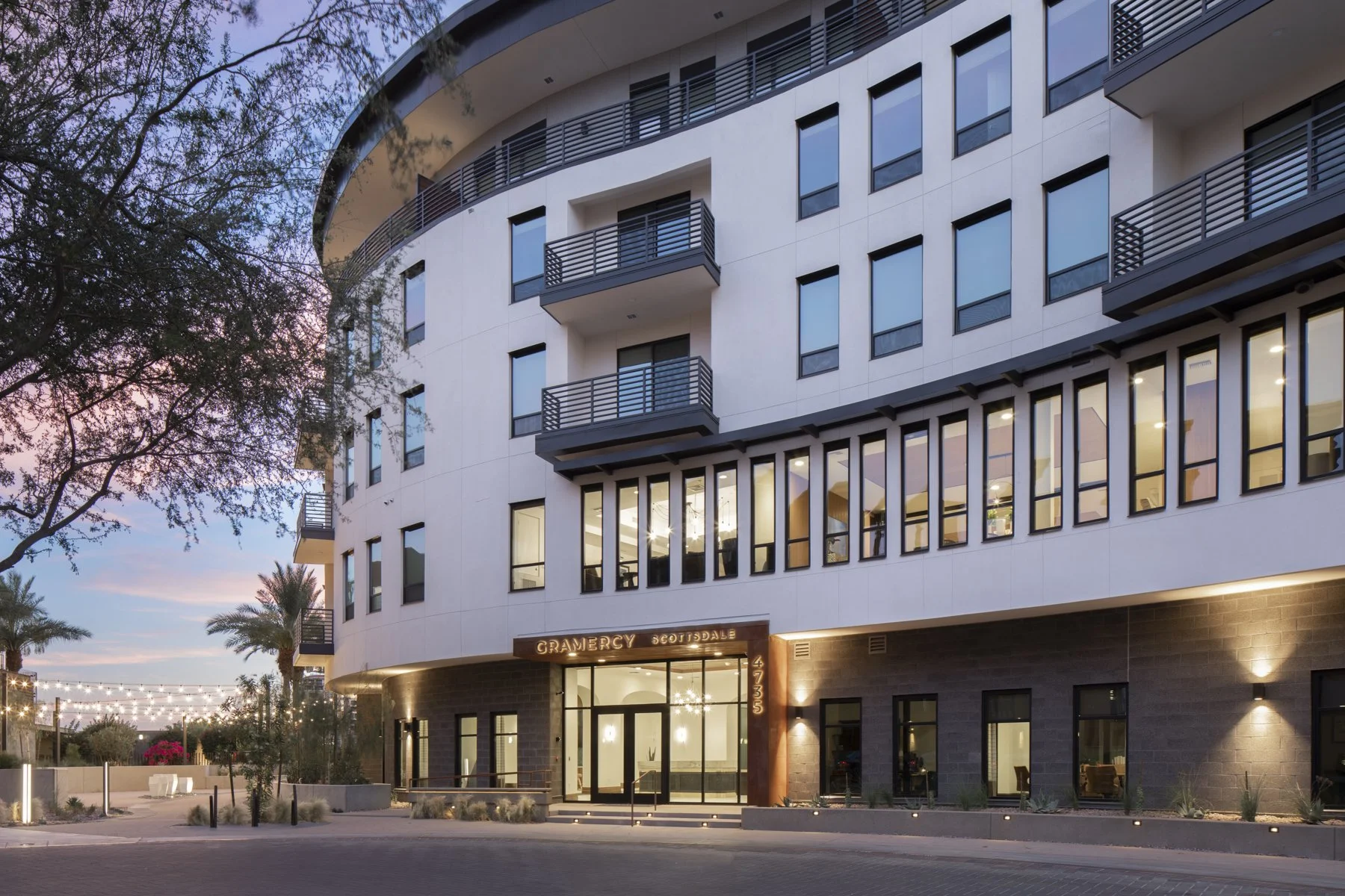 Modern multi-story building with curved design, large glass windows, and balconies. The building has a sign that reads "GRAMERCY SCOTTSDALE" above the entrance, with palm trees and string lights in the surrounding area, suggesting an upscale residential or hotel development in a warm climate.