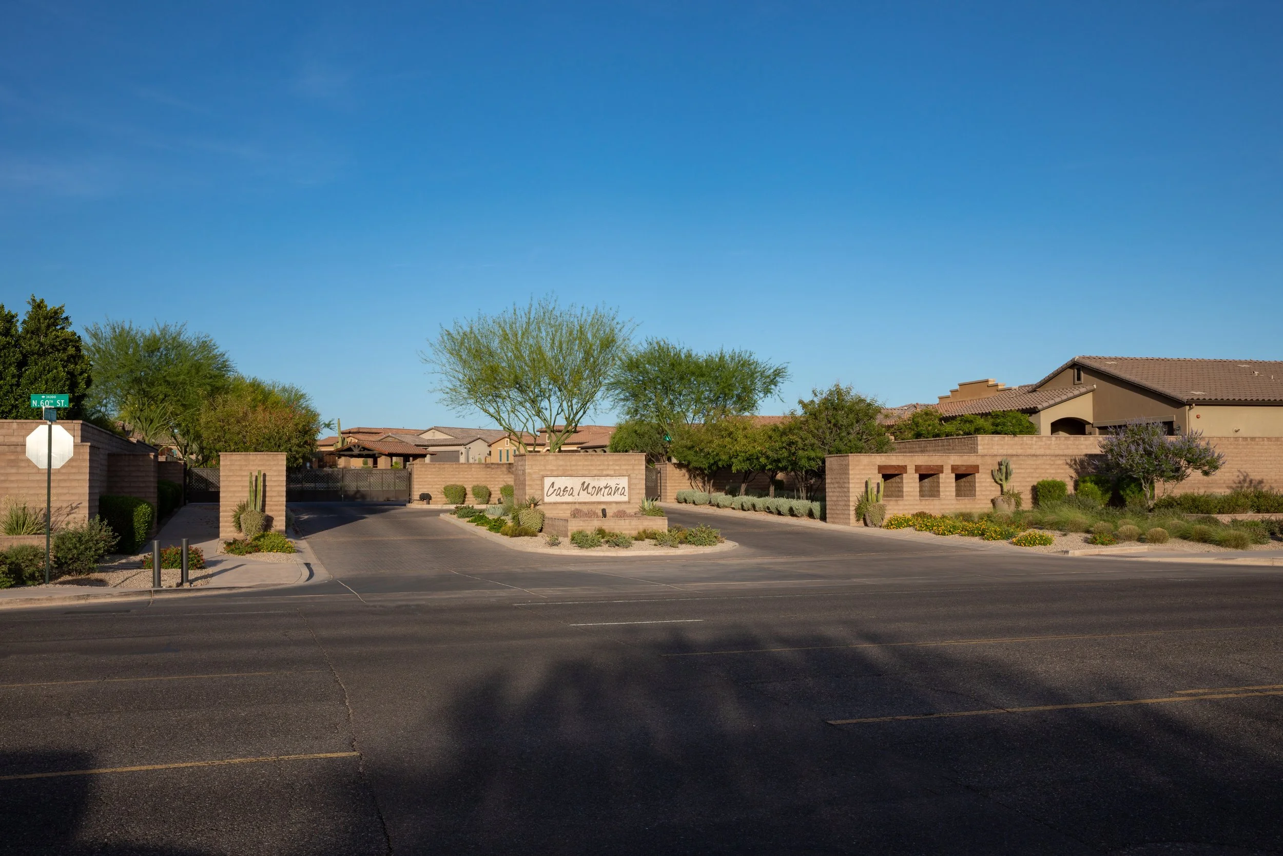 View of a residential neighborhood entrance with a sign that reads 'Casa Montana,' surrounded by desert-style landscaping, trees, and cacti under a clear blue sky.