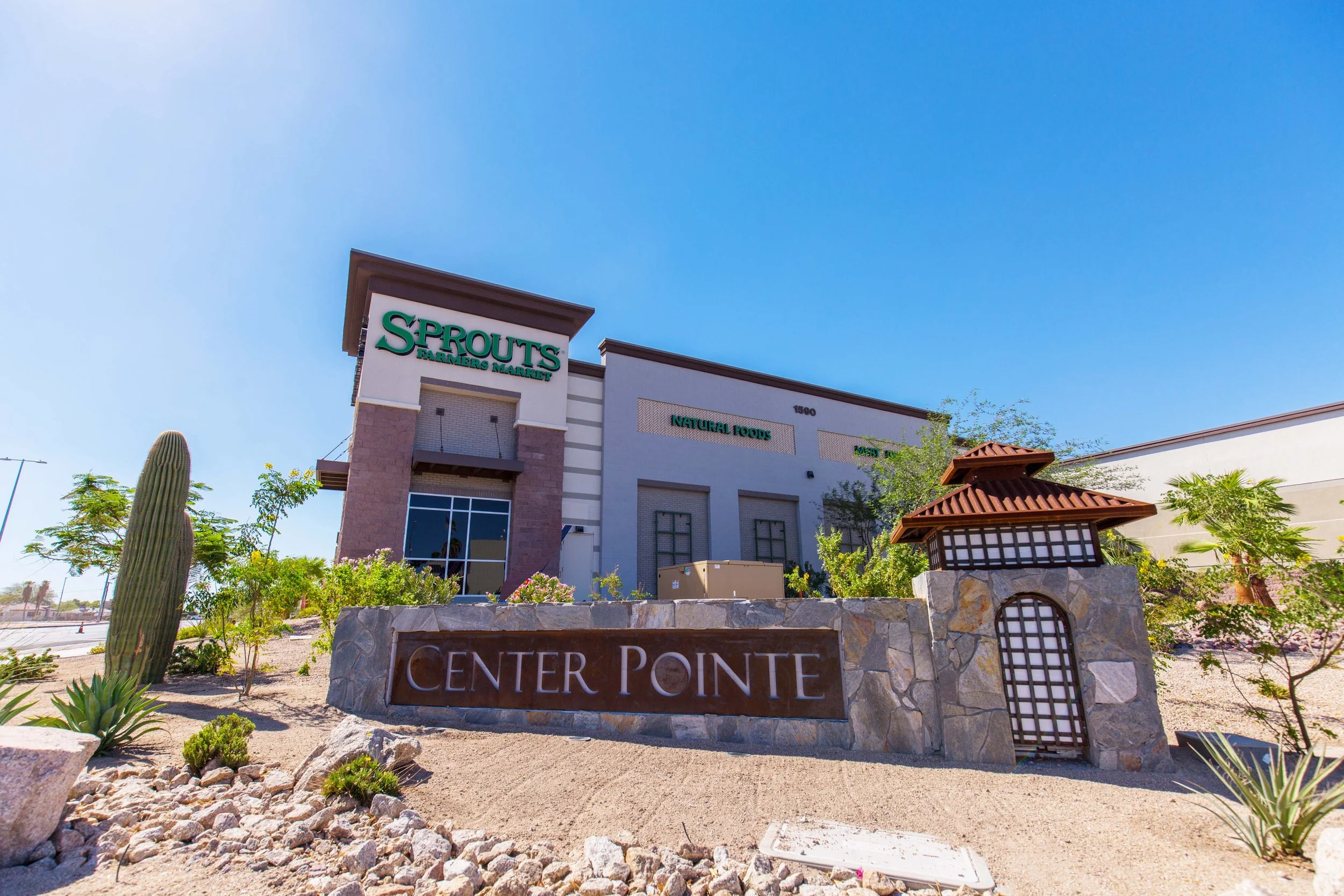 Outside view of a Sprouts Farmers Market store with blue sky overhead, desert landscaping, and a stone sign that reads 'Center Point' in front of the store.