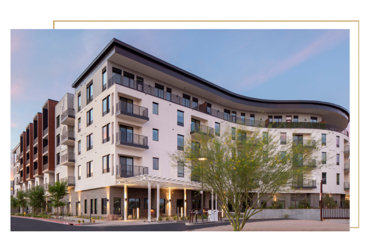Modern multi-story apartment building with balconies, situated at sunset, with trees and a decorative entrance canopy.