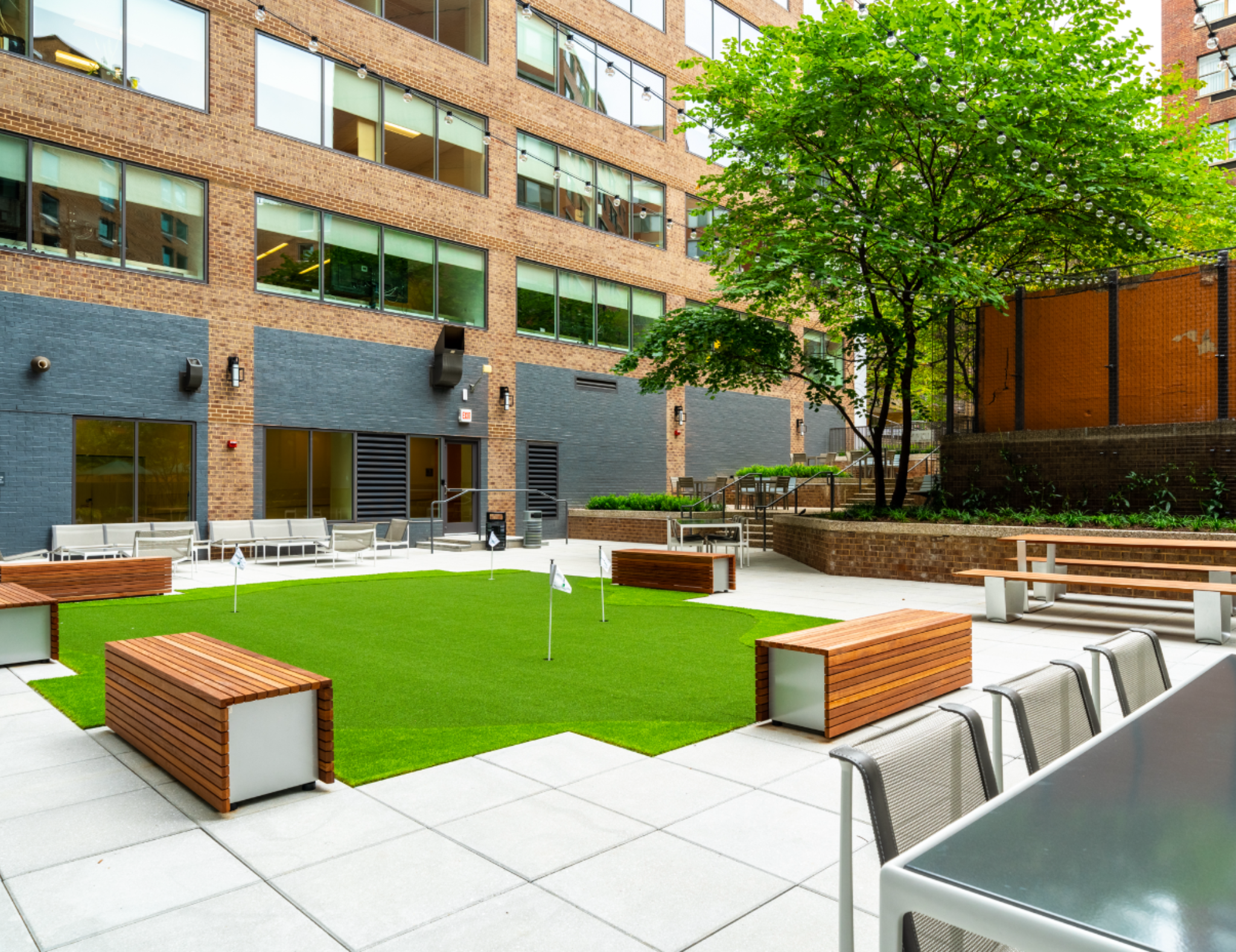 An outdoor courtyard with a green lawn, several benches, and seating areas with metal chairs. There are large brick and glass residential or commercial buildings in the background. A large leafy tree and string lights are also visible.