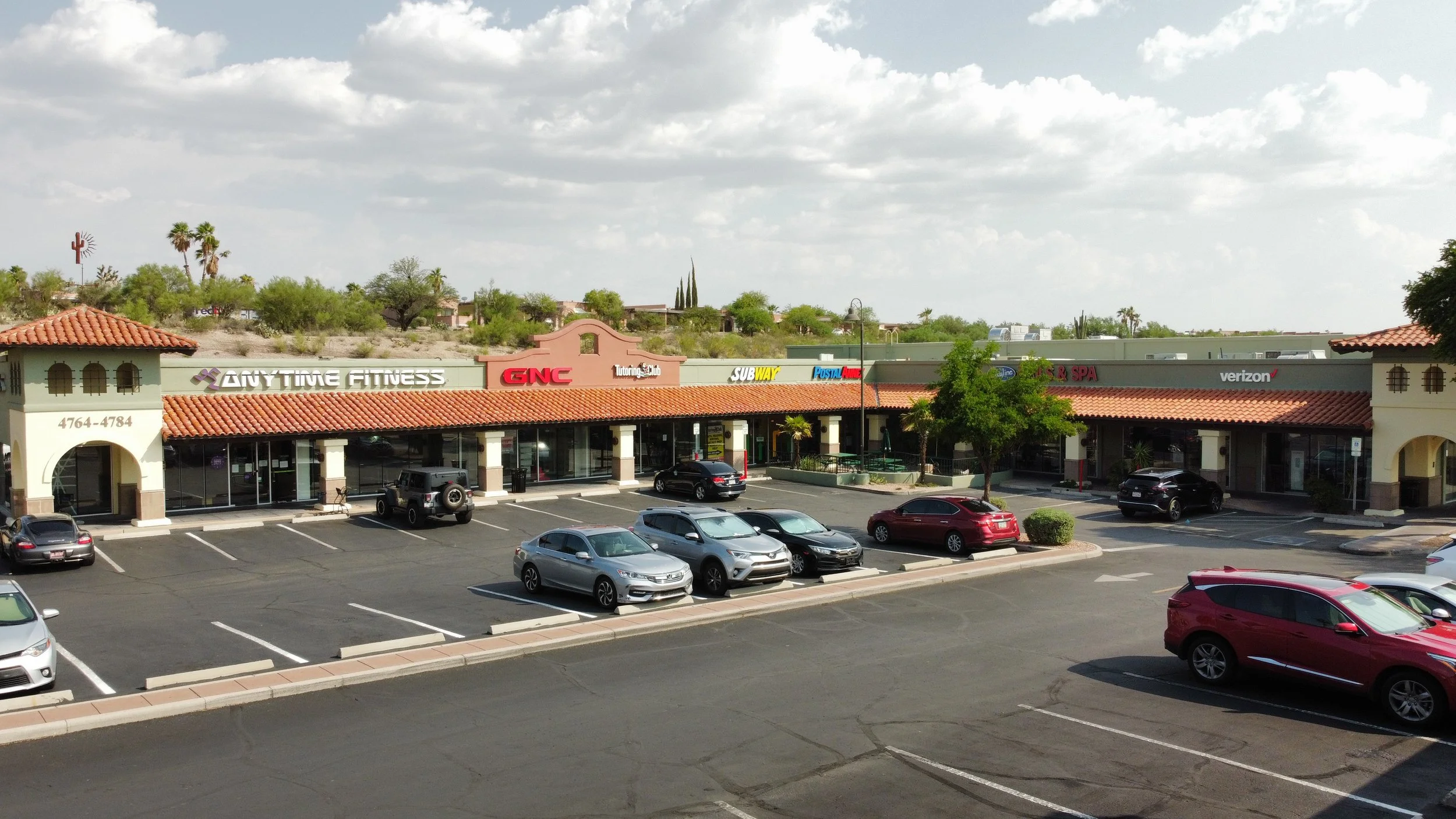 A shopping plaza with various stores and restaurants, including Anytime Fitness, GNC, Subway, PostalAnnex, and Verizon, with cars parked in front and a partly cloudy sky above.