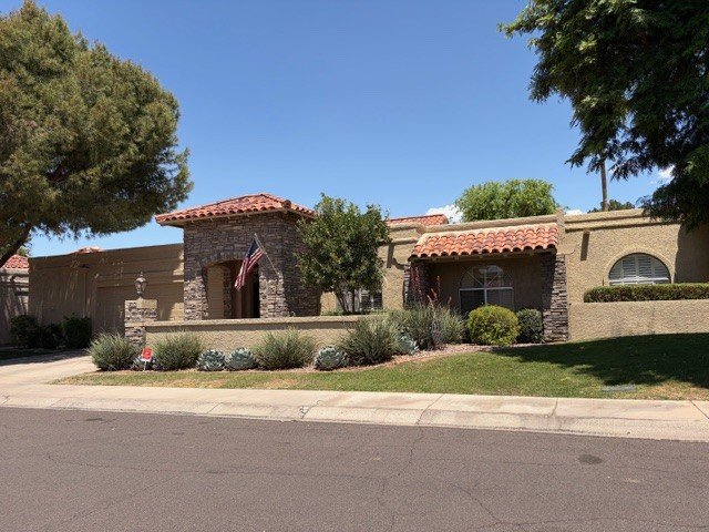 Front view of a residential house with stone accents and a tiled roof, surrounded by trees and landscaping, under a clear blue sky.