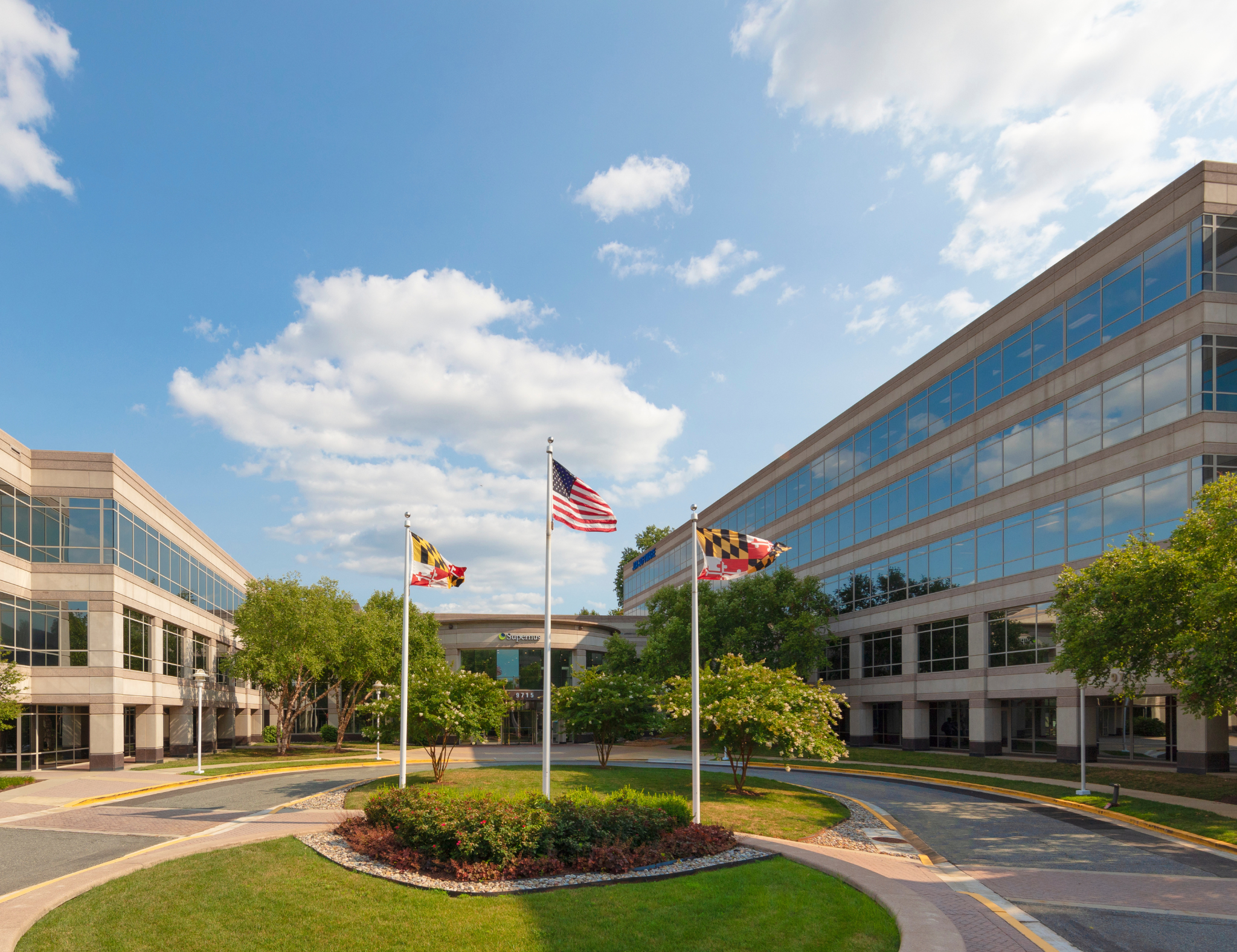 A modern multi-story office building with glass windows, surrounded by green trees, with a lawn and a flower bed in the foreground. Several flags, including the United States flag, are on poles in front of the building, under a partly cloudy blue sky.