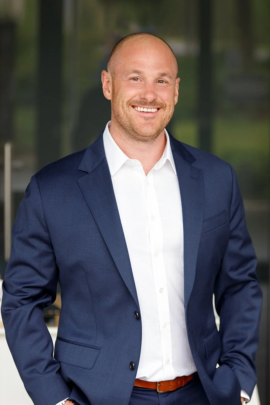 Portrait of a smiling man in a navy suit and white shirt, standing indoors with a blurred glass background.