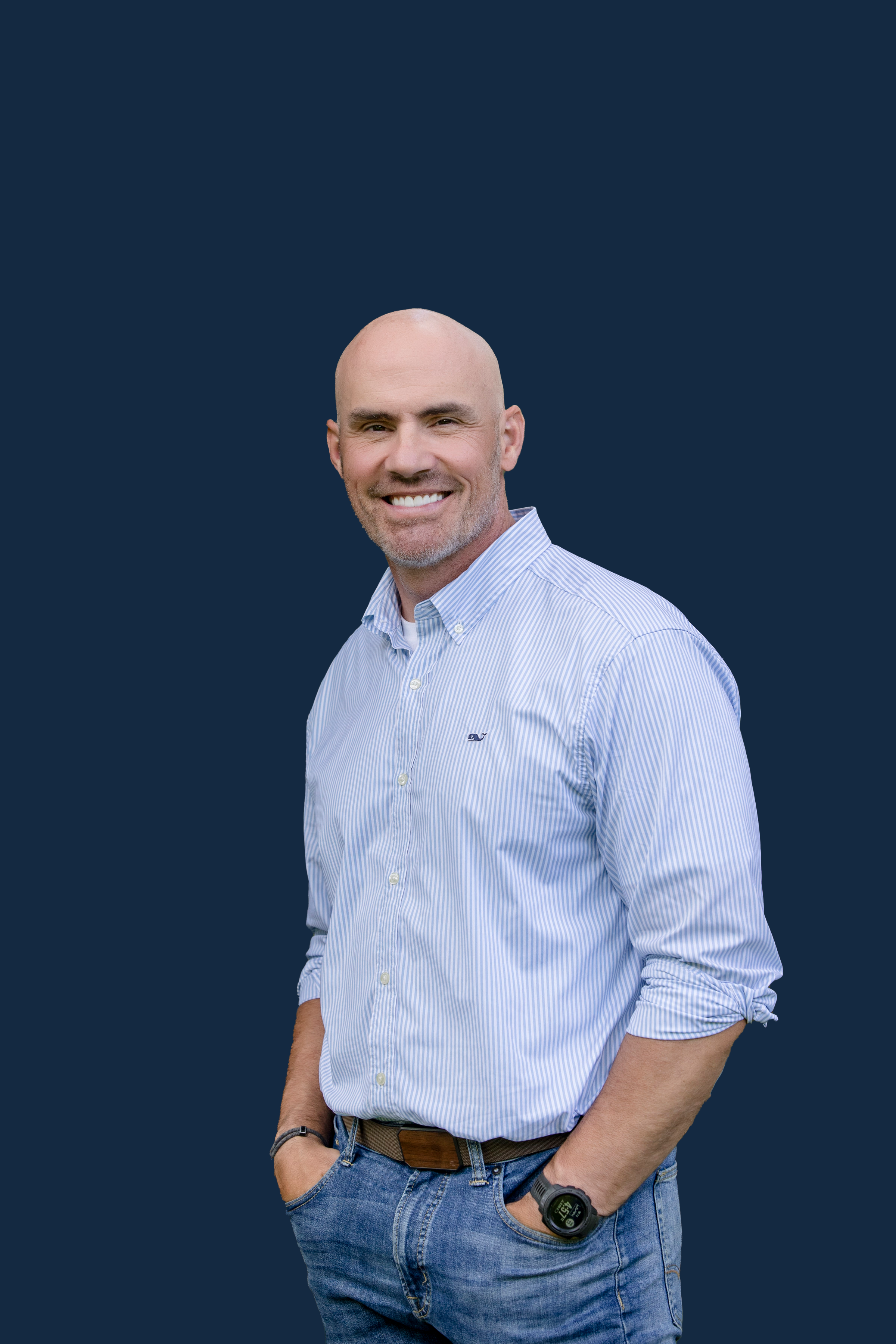 A smiling bald man with a gray beard wearing a light blue striped button-up shirt and jeans, standing with his right hand in his pocket against a dark blue background.