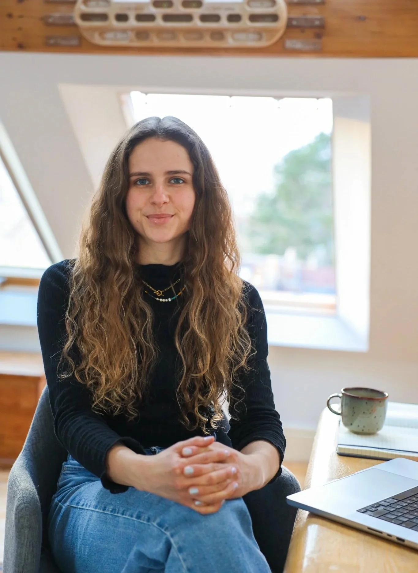 Young woman with long wavy hair sitting at a table with a mug and a laptop, in a room with large windows and a wooden ceiling.