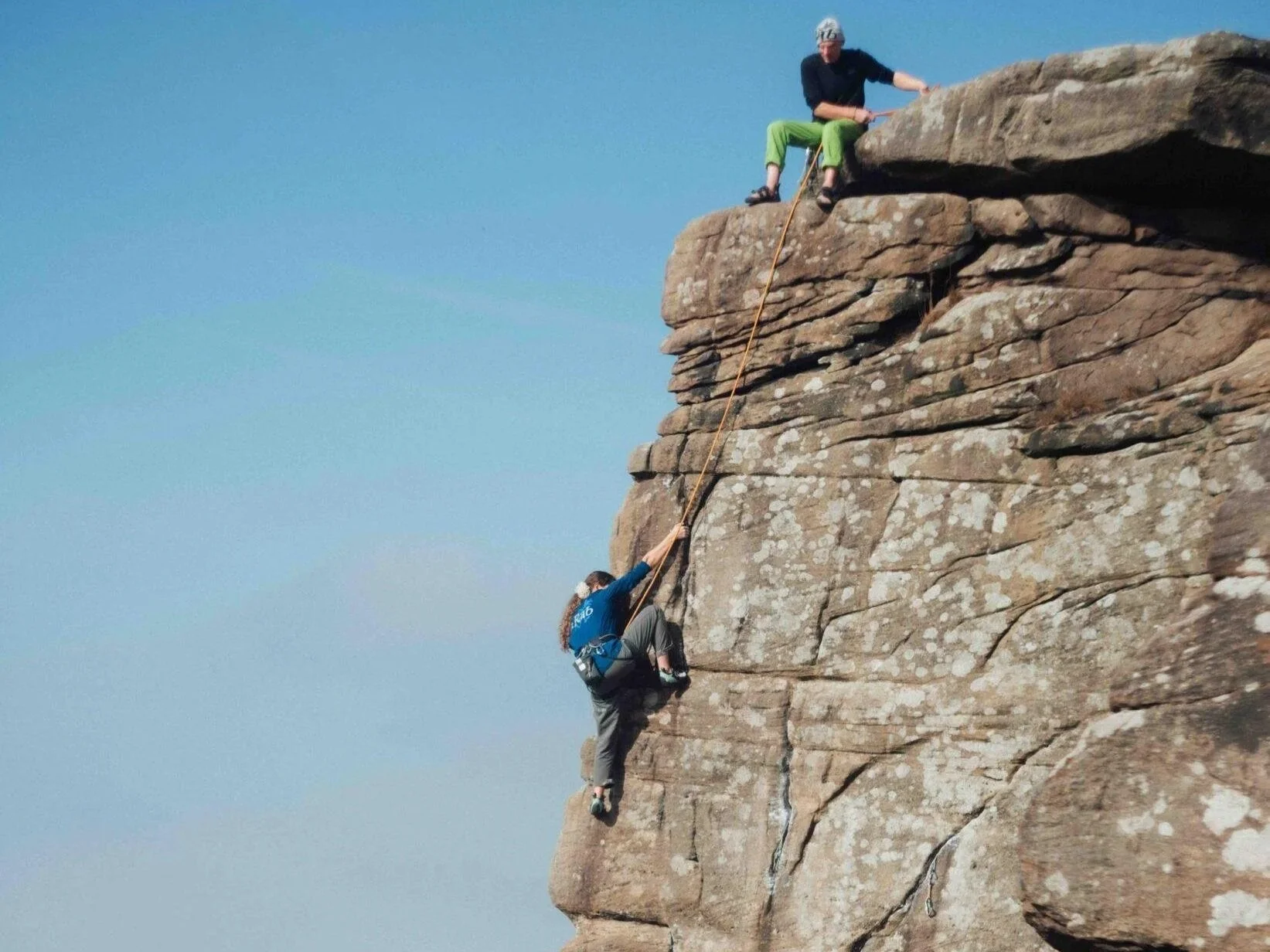 Two people climbing a rocky cliff with one person at the top and the other person climbing up. The climbers are secured with ropes and safety gear.